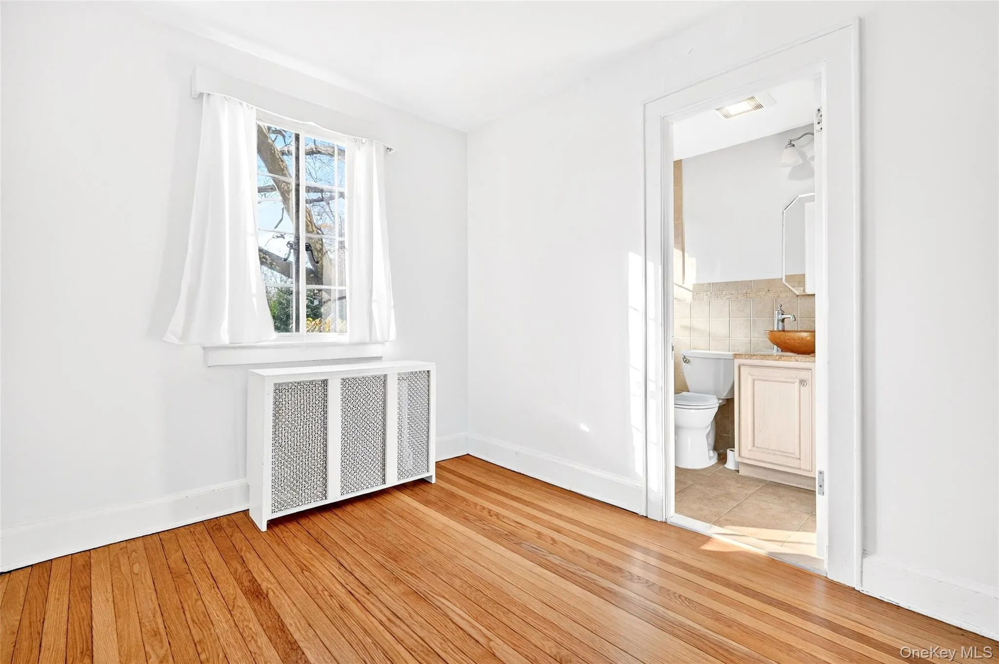 Unfurnished bedroom with radiator, light wood finished floors, and connected bathroom Unfurnished bedroom with radiator, light wood finished floors, and connected bathroom