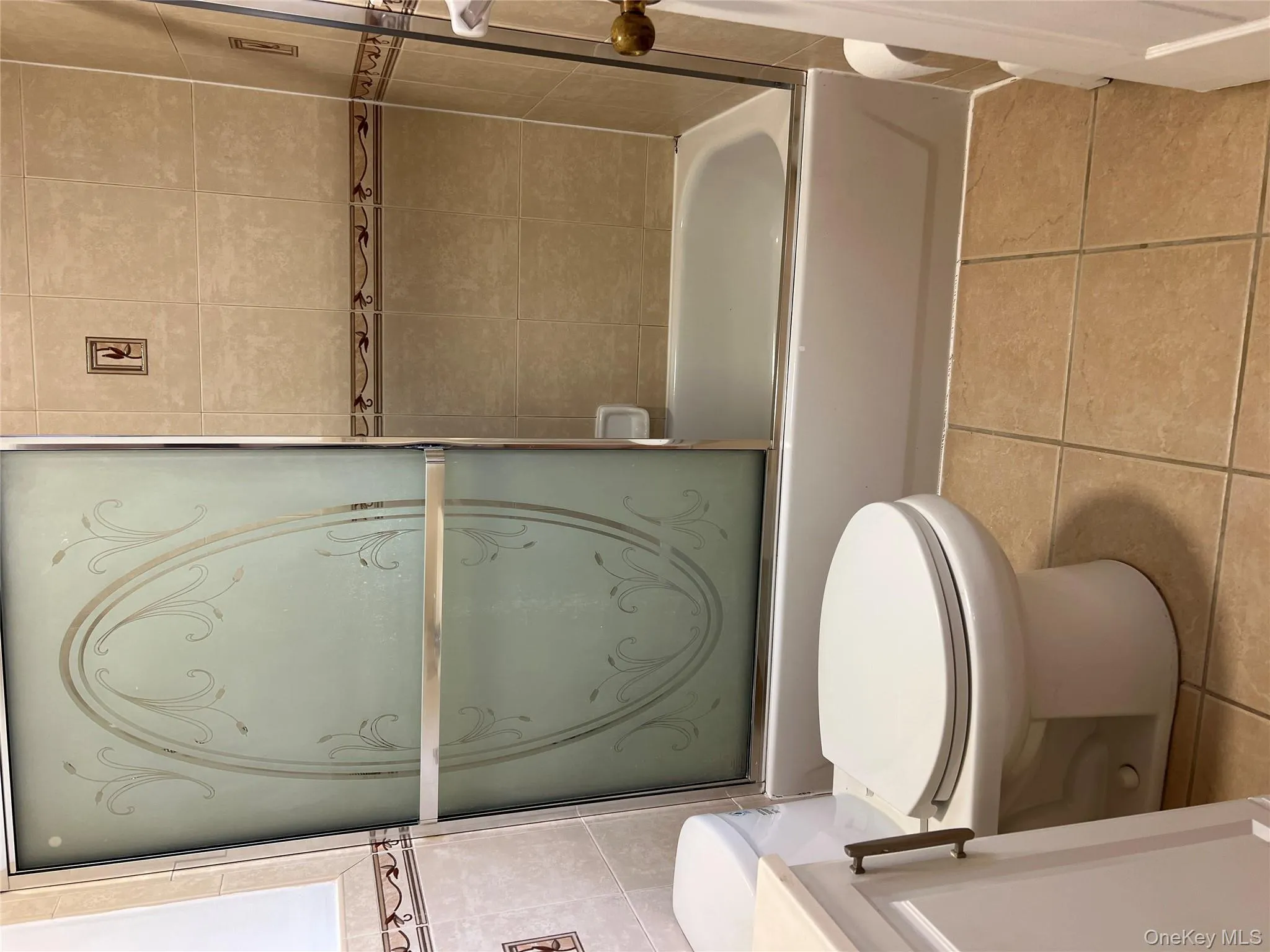 Full bath featuring light tile patterned flooring, shower / bath combination with glass door, tile walls, and vanity Full bath featuring light tile patterned flooring, shower / bath combination with glass door, tile walls, and vanity