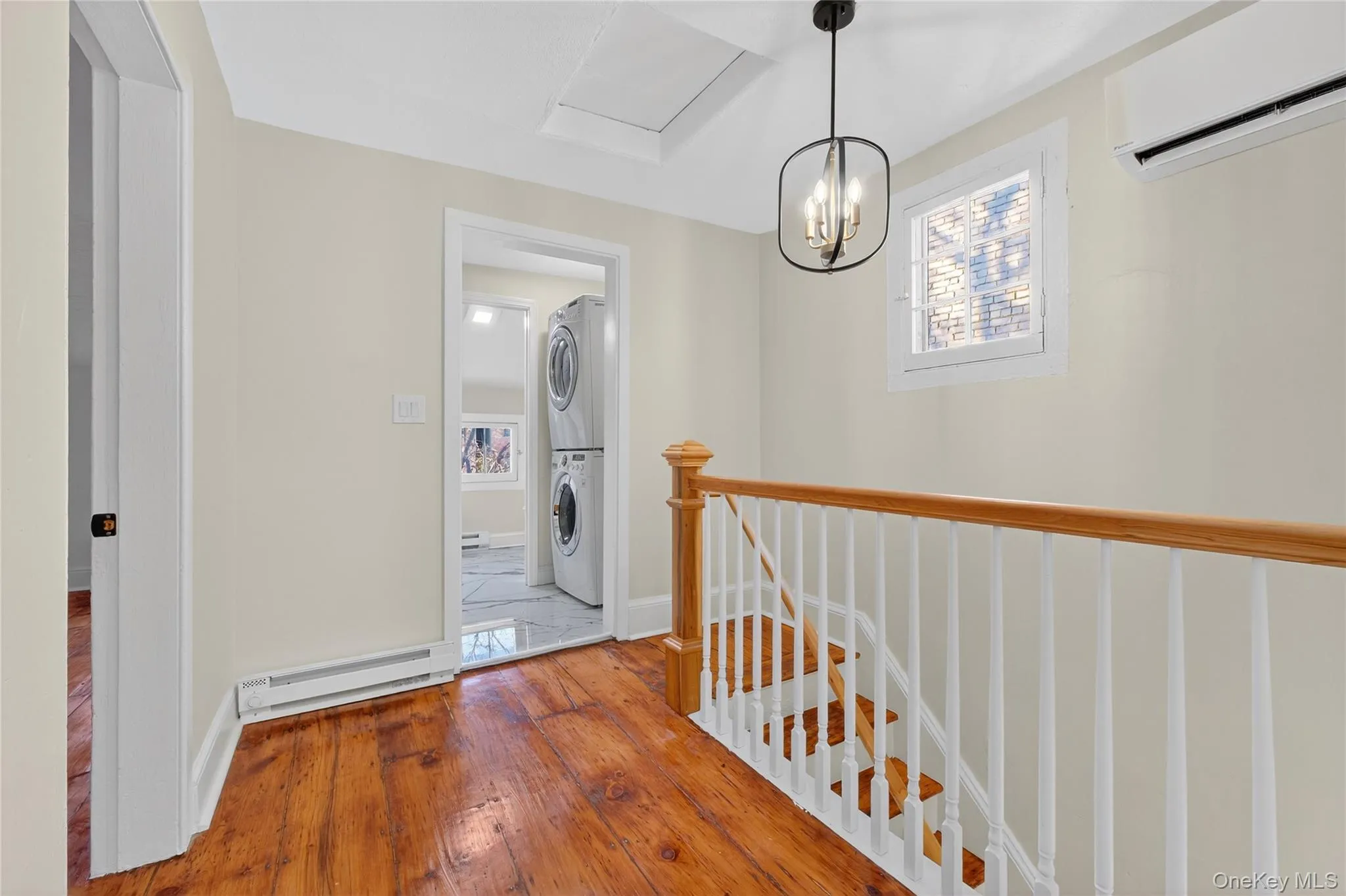 Hall with an upstairs landing, an AC wall unit, a baseboard radiator, stacked washer / drying machine, and hardwood / wood-style floors Hall with an upstairs landing, an AC wall unit, a baseboard radiator, stacked washer / drying machine, and hardwood / wood-style floors
