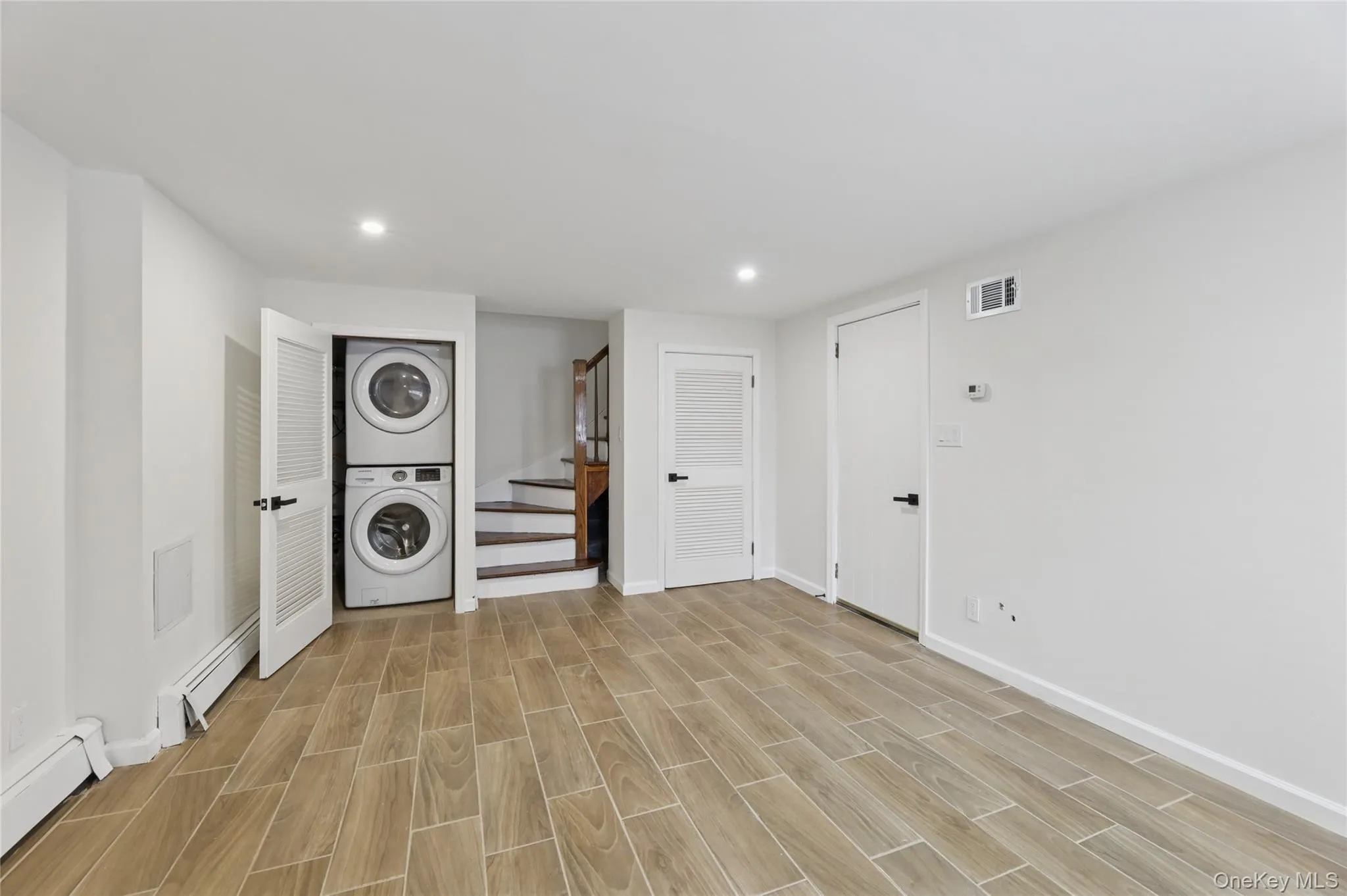 Washroom featuring stacked washer / dryer, wood finish floors, a baseboard heating unit, baseboard heating, and recessed lighting Washroom featuring stacked washer / dryer, wood finish floors, a baseboard heating unit, baseboard heating, and recessed lighting