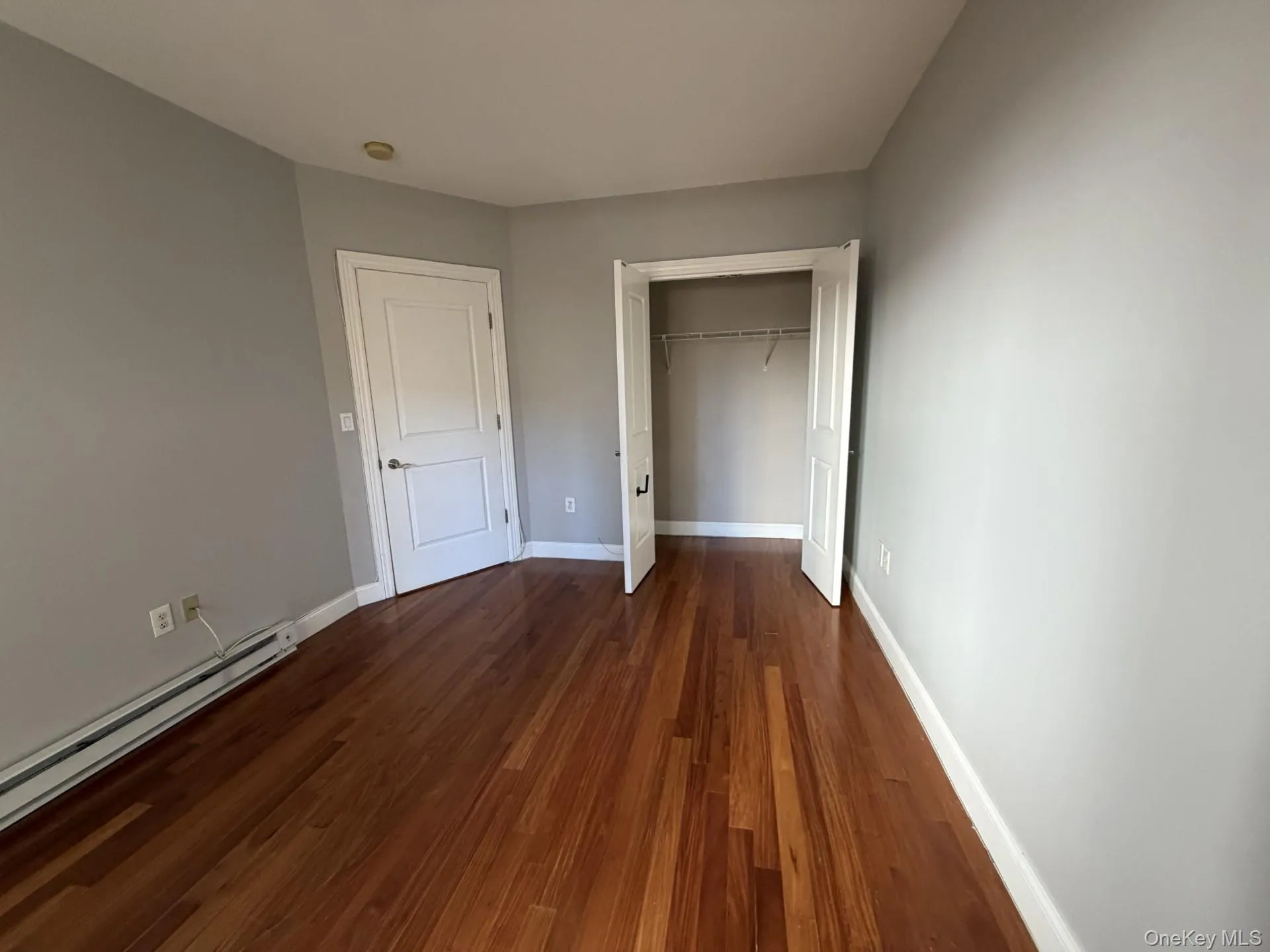 Unfurnished bedroom featuring dark wood-style flooring, a closet, and a baseboard radiator Unfurnished bedroom featuring dark wood-style flooring, a closet, and a baseboard radiator
