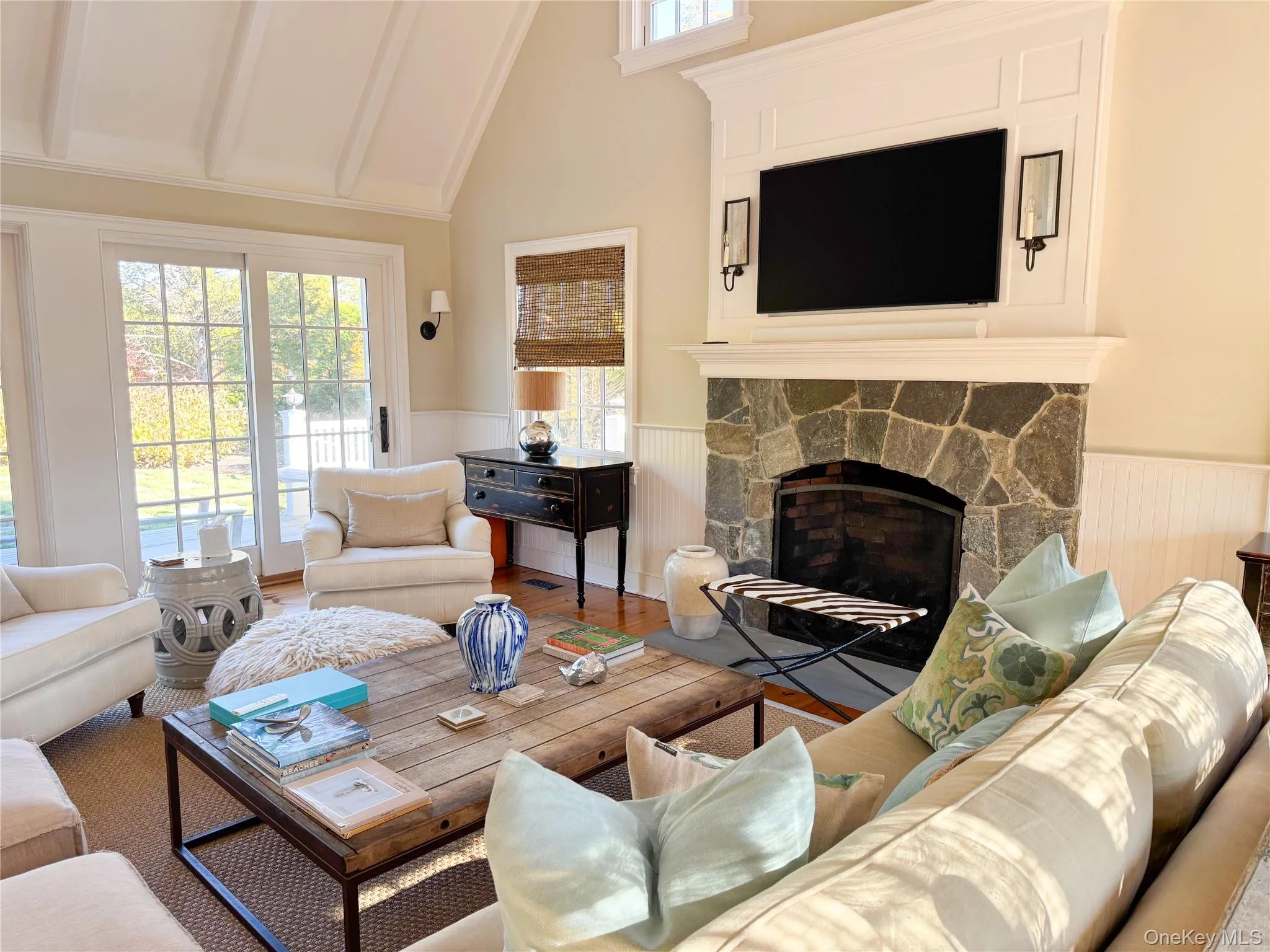 Living area featuring a stone fireplace, wainscoting, beamed ceiling, wood finished floors, and high vaulted ceiling Living area featuring a stone fireplace, wainscoting, beamed ceiling, wood finished floors, and high vaulted ceiling