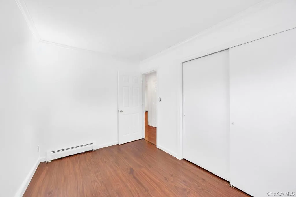 Unfurnished bedroom with baseboard heating, dark wood-type flooring, ornamental molding, and a closet Unfurnished bedroom with baseboard heating, dark wood-type flooring, ornamental molding, and a closet