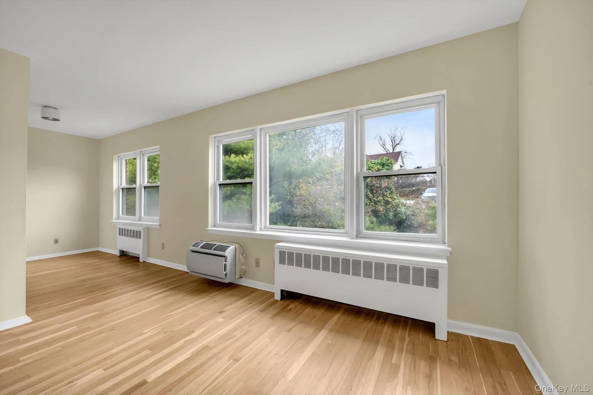 Empty room with radiator, light wood finished floors, and an AC wall unit Empty room with radiator, light wood finished floors, and an AC wall unit