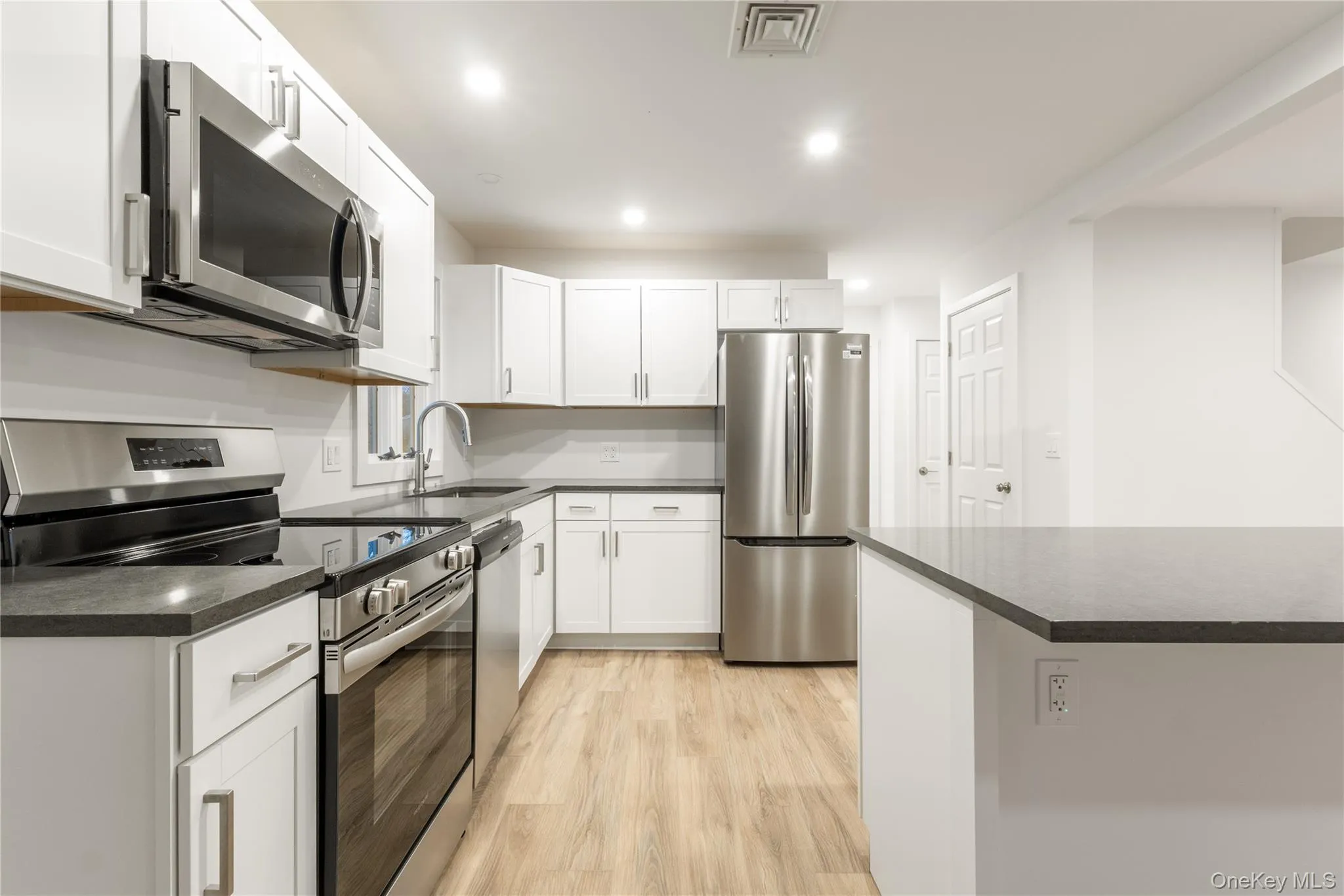 Kitchen with appliances with stainless steel finishes, white cabinets, light wood-type flooring, recessed lighting, and dark stone counters Kitchen with appliances with stainless steel finishes, white cabinets, light wood-type flooring, recessed lighting, and dark stone counters