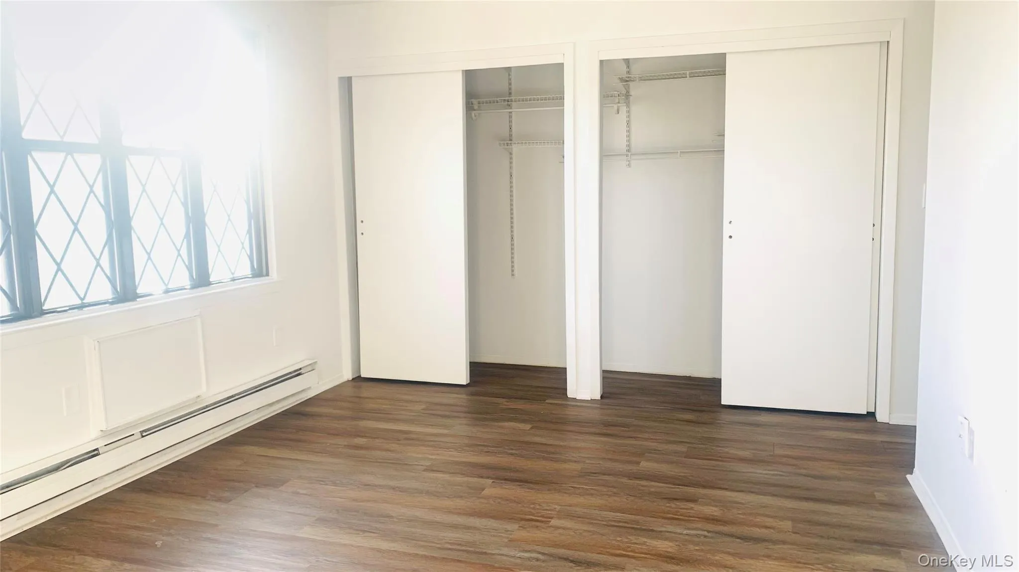 Unfurnished bedroom with two closets, baseboard heating, and dark wood finished floors Unfurnished bedroom with two closets, baseboard heating, and dark wood finished floors