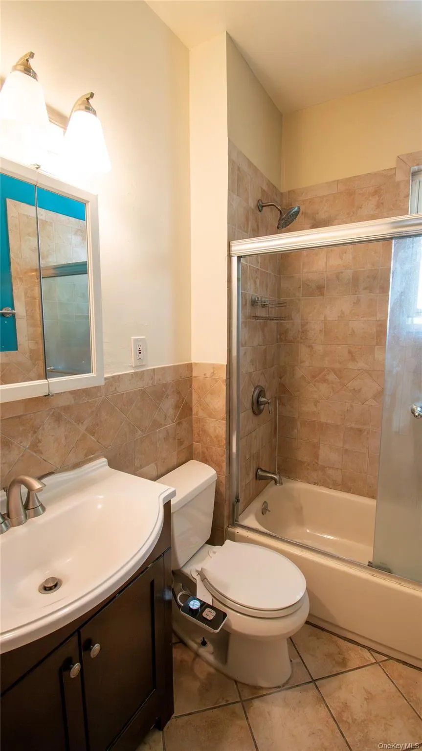 Bathroom featuring vanity, combined bath / shower with glass door, tile walls, light tile patterned floors, and a wainscoted wall Bathroom featuring vanity, combined bath / shower with glass door, tile walls, light tile patterned floors, and a wainscoted wall