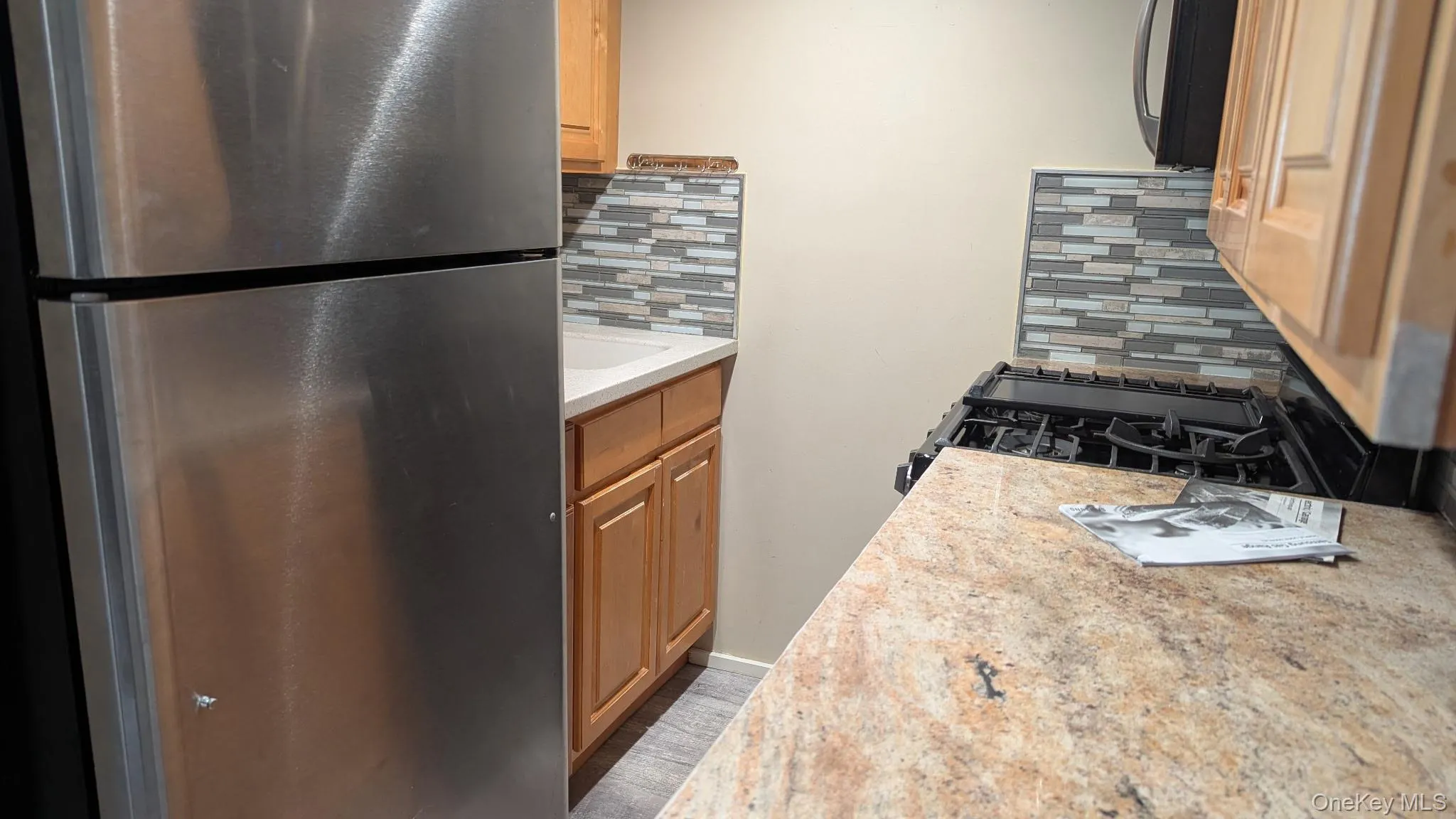 Kitchen with freestanding refrigerator, decorative backsplash, black gas stove, light stone counters, and brown cabinetry Kitchen with freestanding refrigerator, decorative backsplash, black gas stove, light stone counters, and brown cabinetry