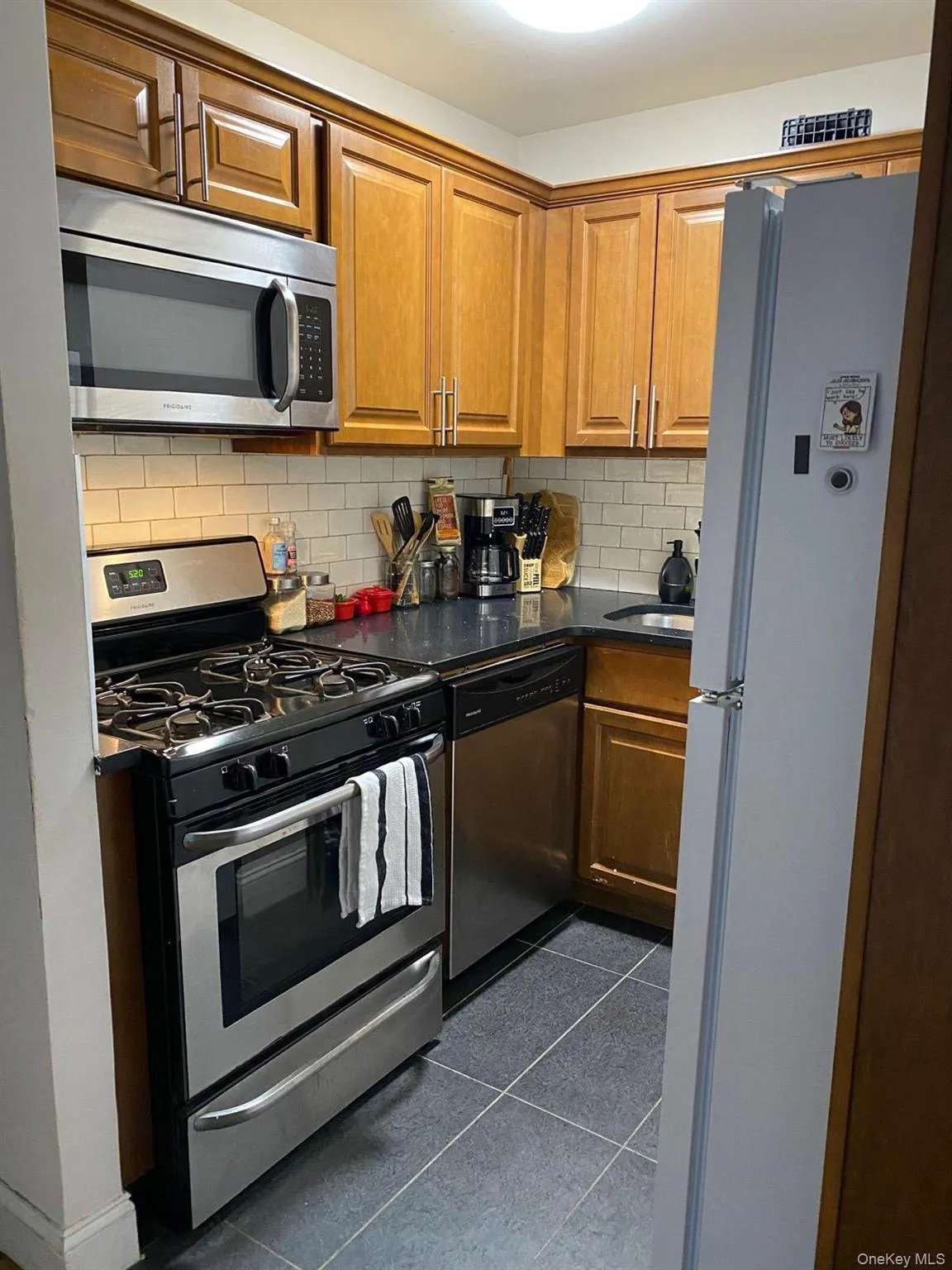 Kitchen with appliances with stainless steel finishes, brown cabinets, decorative backsplash, dark tile patterned flooring, and dark stone countertops Kitchen with appliances with stainless steel finishes, brown cabinets, decorative backsplash, dark tile patterned flooring, and dark stone countertops