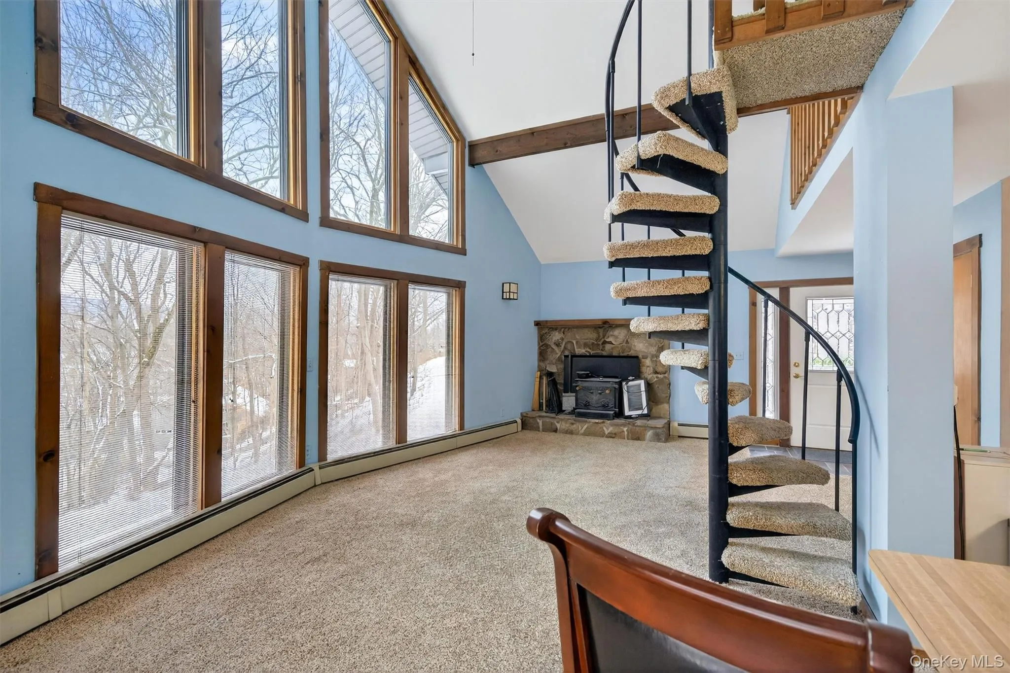 Carpeted living room with baseboard heating, high vaulted ceiling, beamed ceiling, a stone fireplace, and stairway Carpeted living room with baseboard heating, high vaulted ceiling, beamed ceiling, a stone fireplace, and stairway