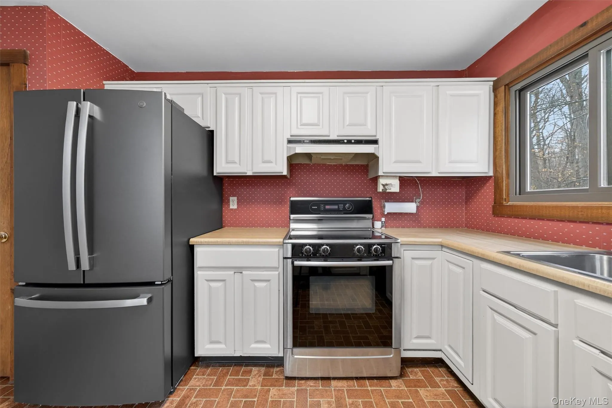 Kitchen featuring appliances with stainless steel finishes, brick patterned flooring, white cabinetry, light countertops, and wallpapered walls Kitchen featuring appliances with stainless steel finishes, brick patterned flooring, white cabinetry, light countertops, and wallpapered walls