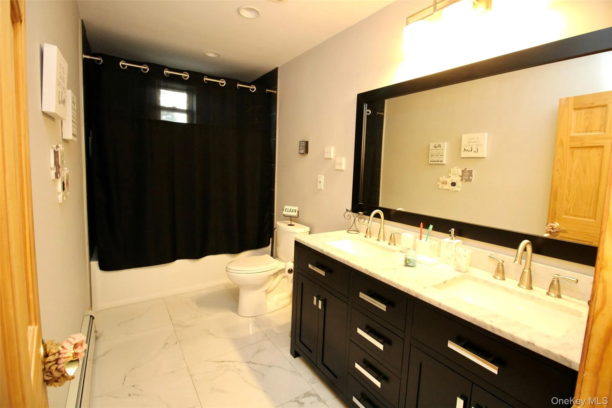 Bathroom with double vanity, shower / tub combo, light marble finish flooring, and a baseboard radiator Bathroom with double vanity, shower / tub combo, light marble finish flooring, and a baseboard radiator