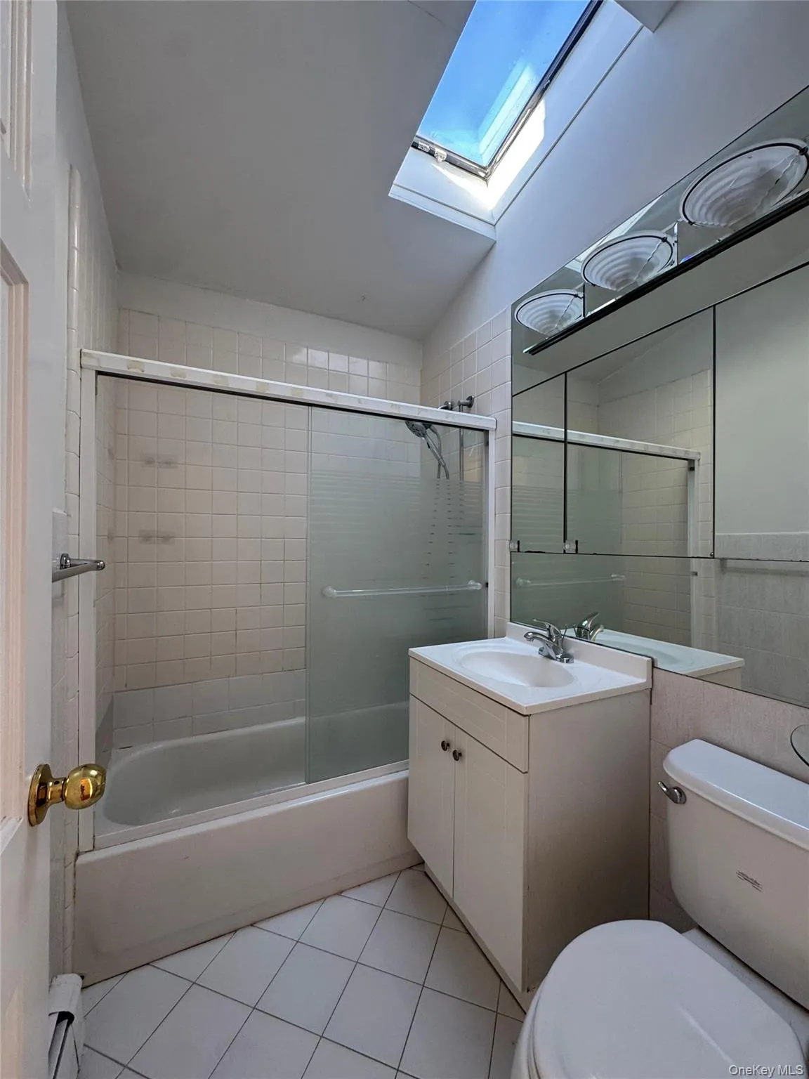 Bathroom featuring light tile patterned floors, bath / shower combo with glass door, vanity, a skylight, and a baseboard radiator Bathroom featuring light tile patterned floors, bath / shower combo with glass door, vanity, a skylight, and a baseboard radiator