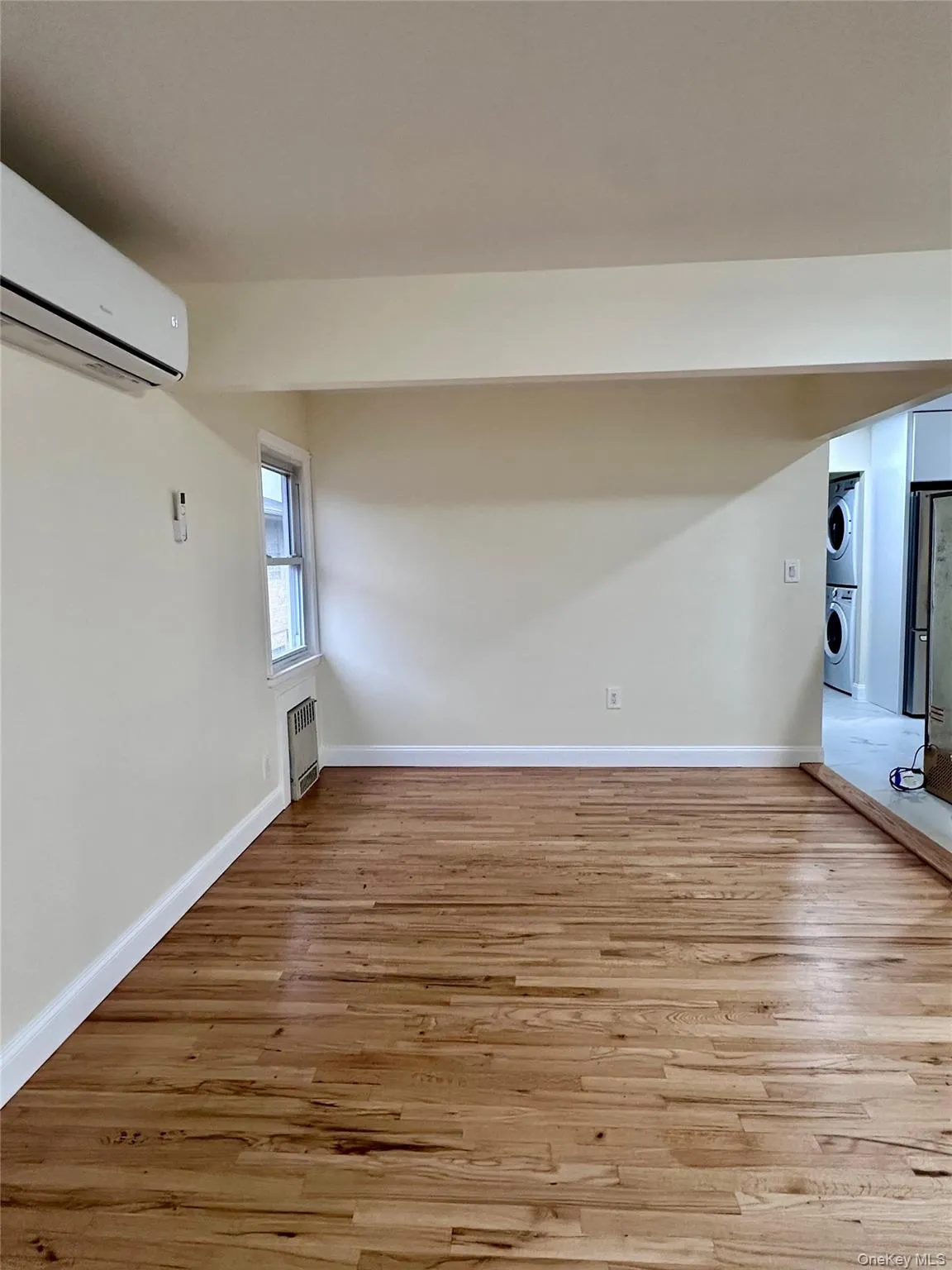 Unfurnished living room with light wood finished floors, stacked washing machine and dryer, and an AC wall unit Unfurnished living room with light wood finished floors, stacked washing machine and dryer, and an AC wall unit
