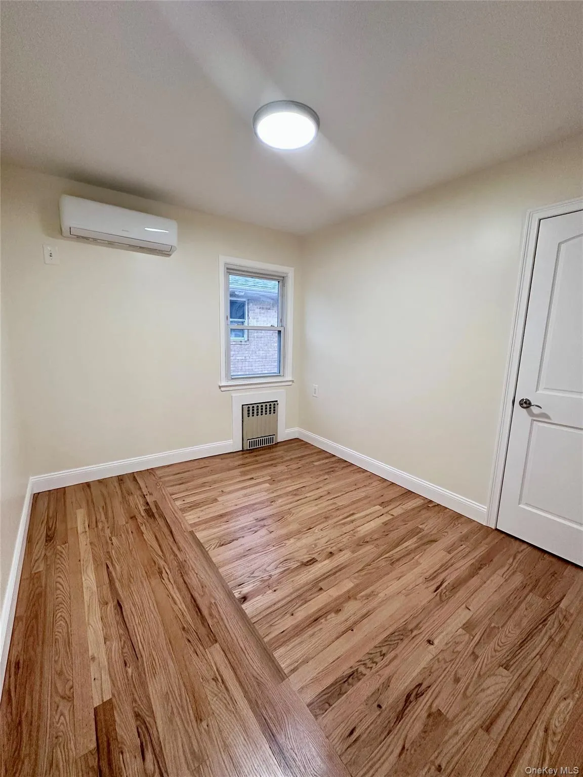 Unfurnished room with light wood finished floors, radiator heating unit, and a wall mounted air conditioner Unfurnished room with light wood finished floors, radiator heating unit, and a wall mounted air conditioner