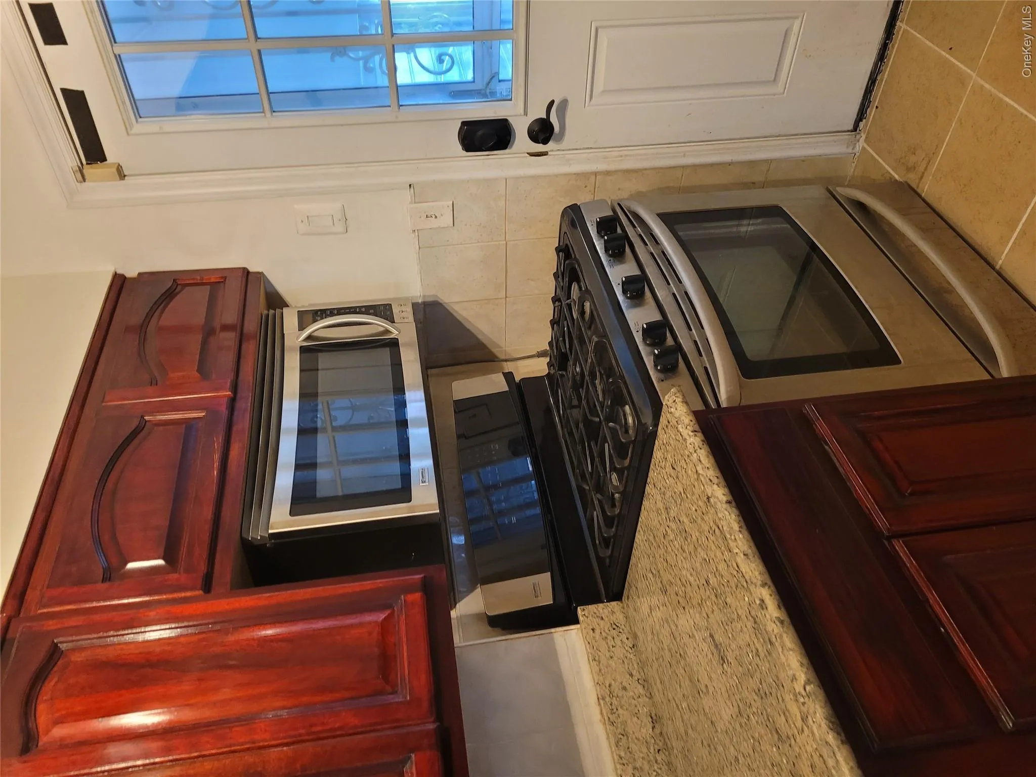 Kitchen featuring reddish brown cabinets, stainless steel range with gas stovetop, light stone countertops, and tile walls Kitchen featuring reddish brown cabinets, stainless steel range with gas stovetop, light stone countertops, and tile walls