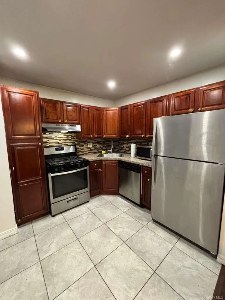 Kitchen with appliances with stainless steel finishes, under cabinet range hood, decorative backsplash, reddish brown cabinets, and recessed lighting Kitchen with appliances with stainless steel finishes, under cabinet range hood, decorative backsplash, reddish brown cabinets, and recessed lighting