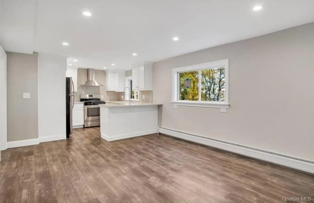 Kitchen featuring a baseboard heating unit, kitchen peninsula, wall chimney exhaust hood, appliances with stainless steel finishes, and white cabinetry Kitchen featuring a baseboard heating unit, kitchen peninsula, wall chimney exhaust hood, appliances with stainless steel finishes, and white cabinetry