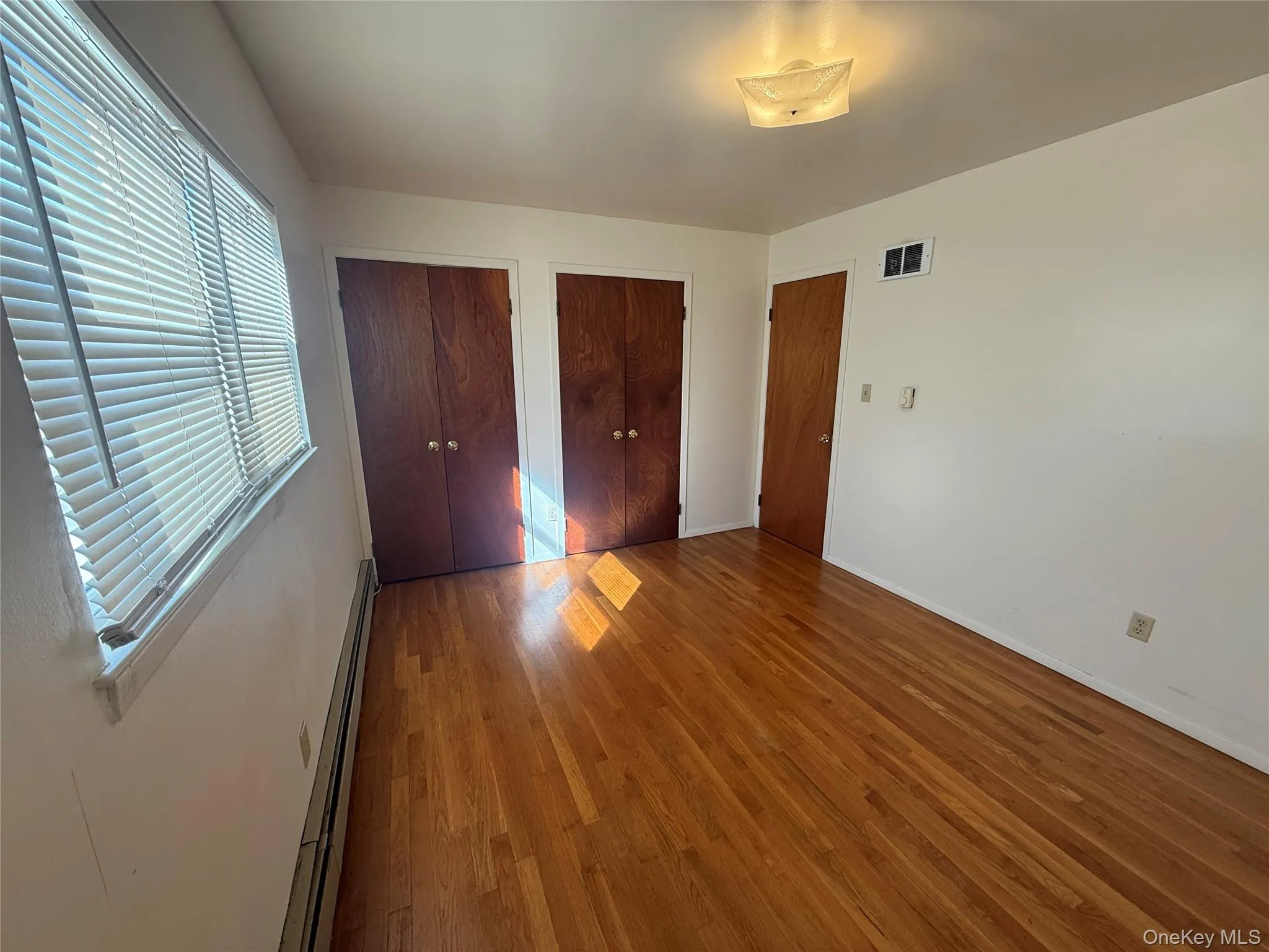 Unfurnished bedroom featuring two closets, wood finished floors, and a baseboard heating unit Unfurnished bedroom featuring two closets, wood finished floors, and a baseboard heating unit