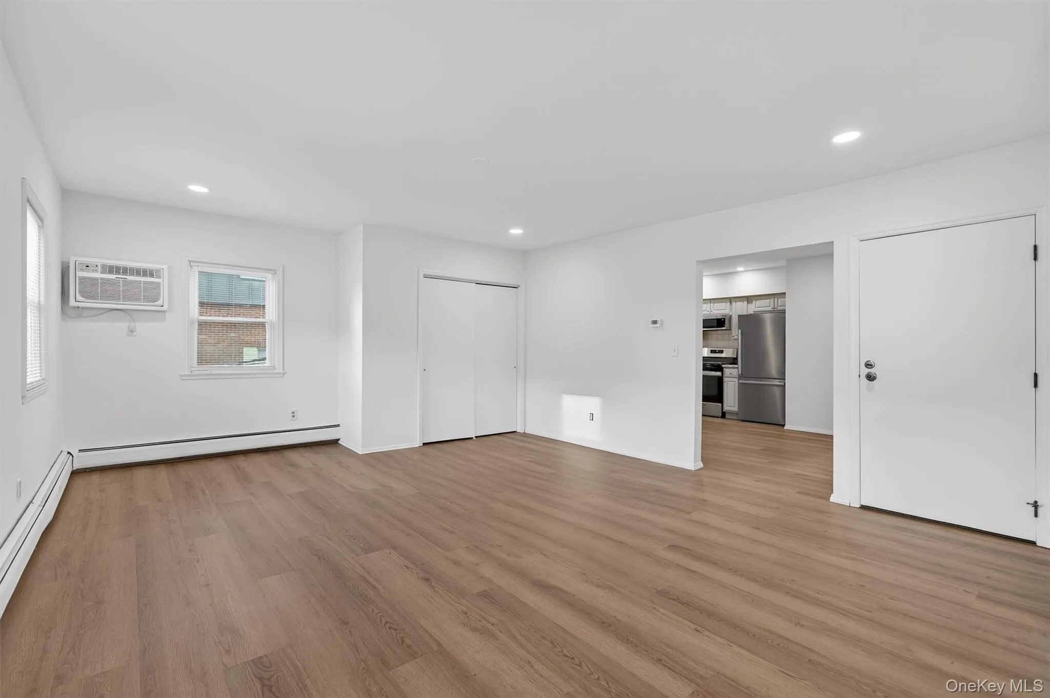 Unfurnished living room with recessed lighting, light wood-style floors, freestanding refrigerator, a baseboard heating unit, and an AC wall unit Unfurnished living room with recessed lighting, light wood-style floors, freestanding refrigerator, a baseboard heating unit, and an AC wall unit