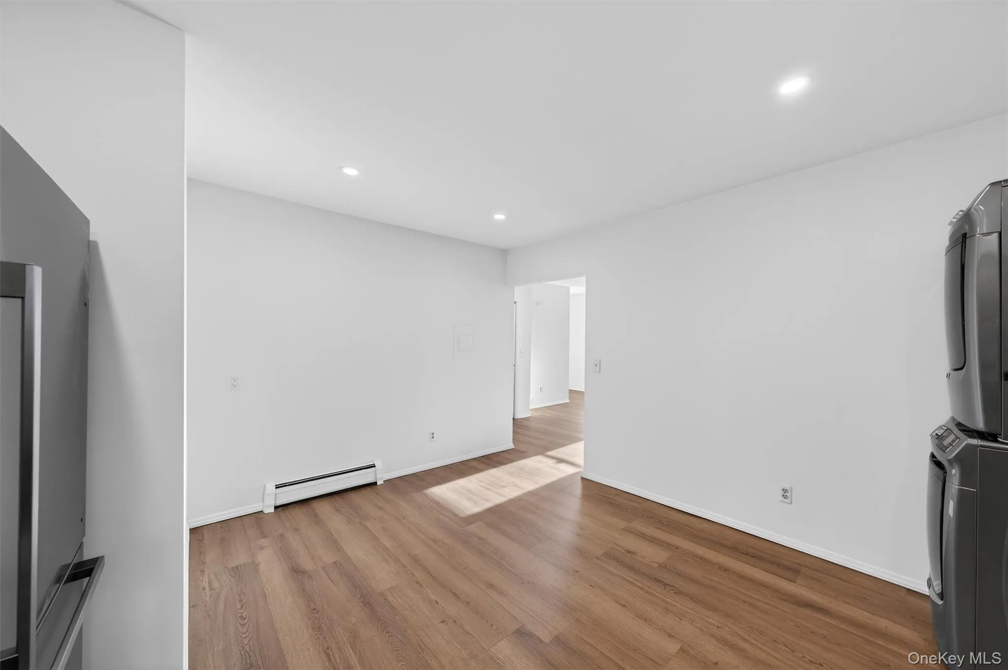 Unfurnished living room with stacked washer / drying machine, a baseboard radiator, light wood-type flooring, and recessed lighting Unfurnished living room with stacked washer / drying machine, a baseboard radiator, light wood-type flooring, and recessed lighting