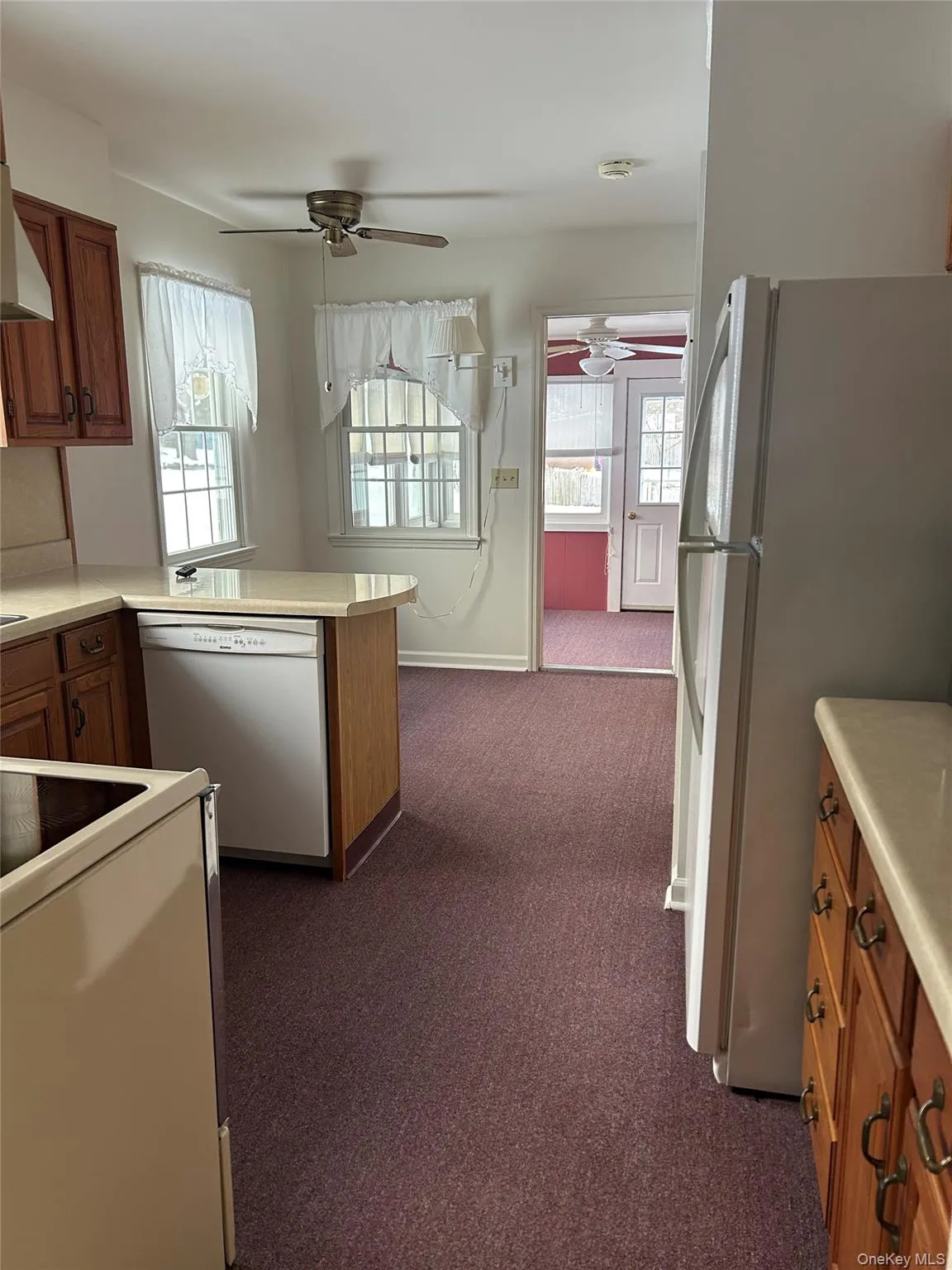 Kitchen featuring brown cabinetry, light countertops, white appliances, dark carpet, and plenty of natural light Kitchen featuring brown cabinetry, light countertops, white appliances, dark carpet, and plenty of natural light