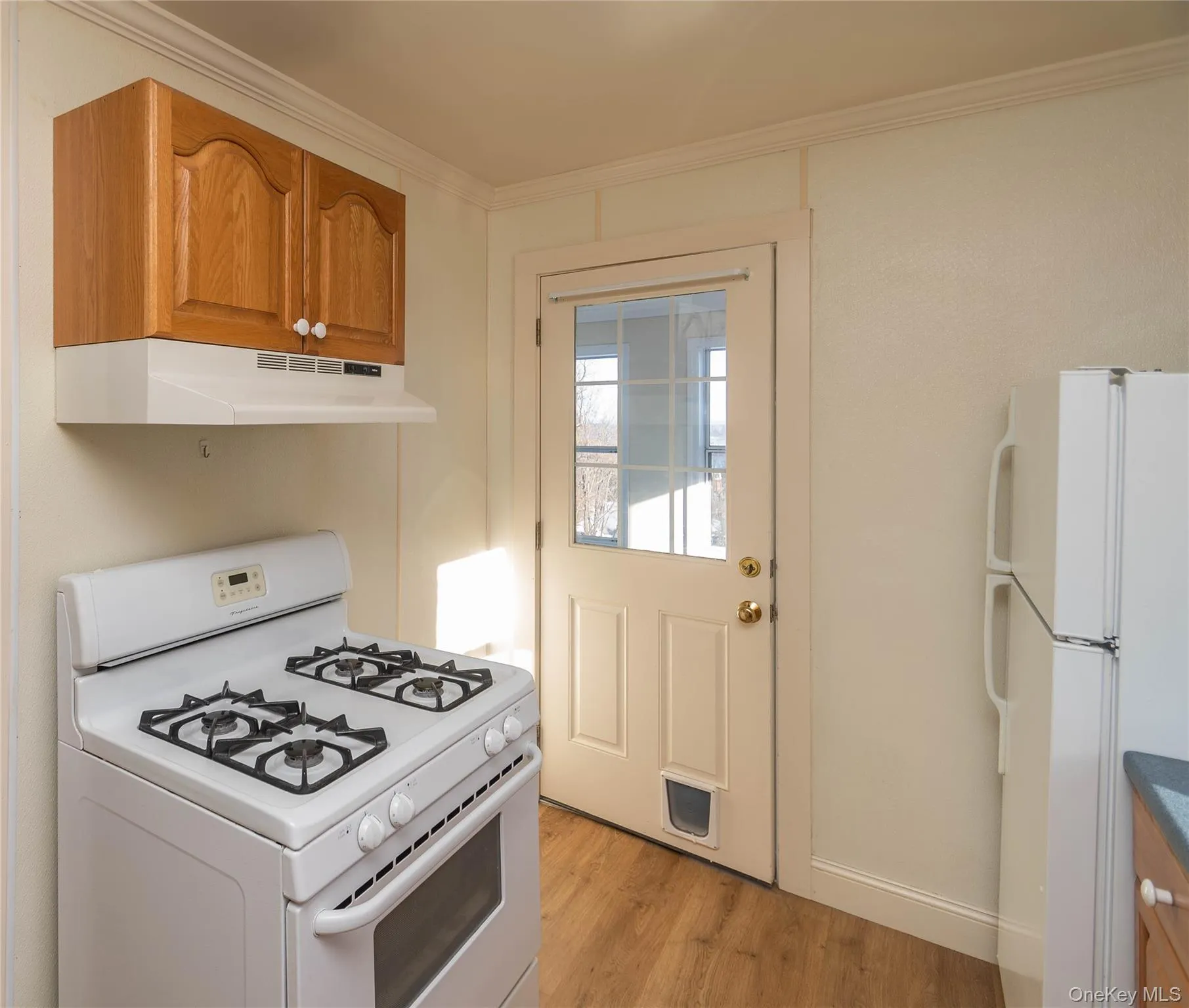Kitchen featuring white appliances, under cabinet range hood, light wood-type flooring, brown cabinets, and ornamental molding Kitchen featuring white appliances, under cabinet range hood, light wood-type flooring, brown cabinets, and ornamental molding