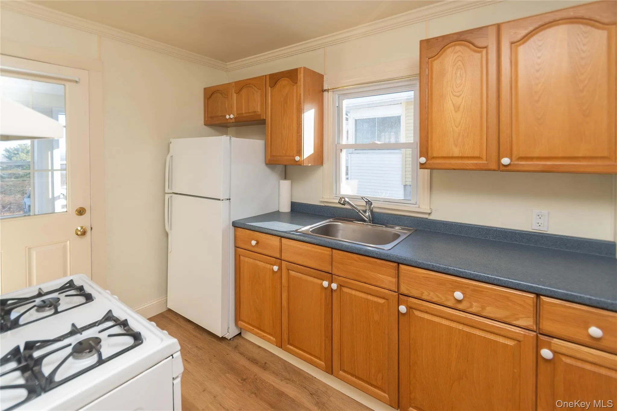 Kitchen featuring white appliances, dark countertops, brown cabinetry, light wood finished floors, and crown molding Kitchen featuring white appliances, dark countertops, brown cabinetry, light wood finished floors, and crown molding