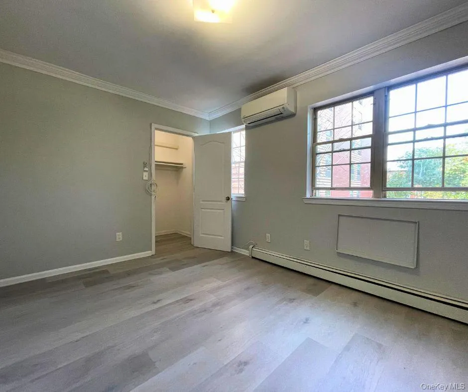 Unfurnished bedroom featuring baseboard heating, a walk in closet, crown molding, light wood-style flooring, and an AC wall unit Unfurnished bedroom featuring baseboard heating, a walk in closet, crown molding, light wood-style flooring, and an AC wall unit
