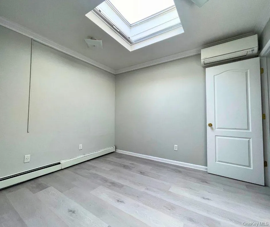 Unfurnished room with a skylight, a wall mounted air conditioner, a baseboard heating unit, light wood-type flooring, and crown molding Unfurnished room with a skylight, a wall mounted air conditioner, a baseboard heating unit, light wood-type flooring, and crown molding