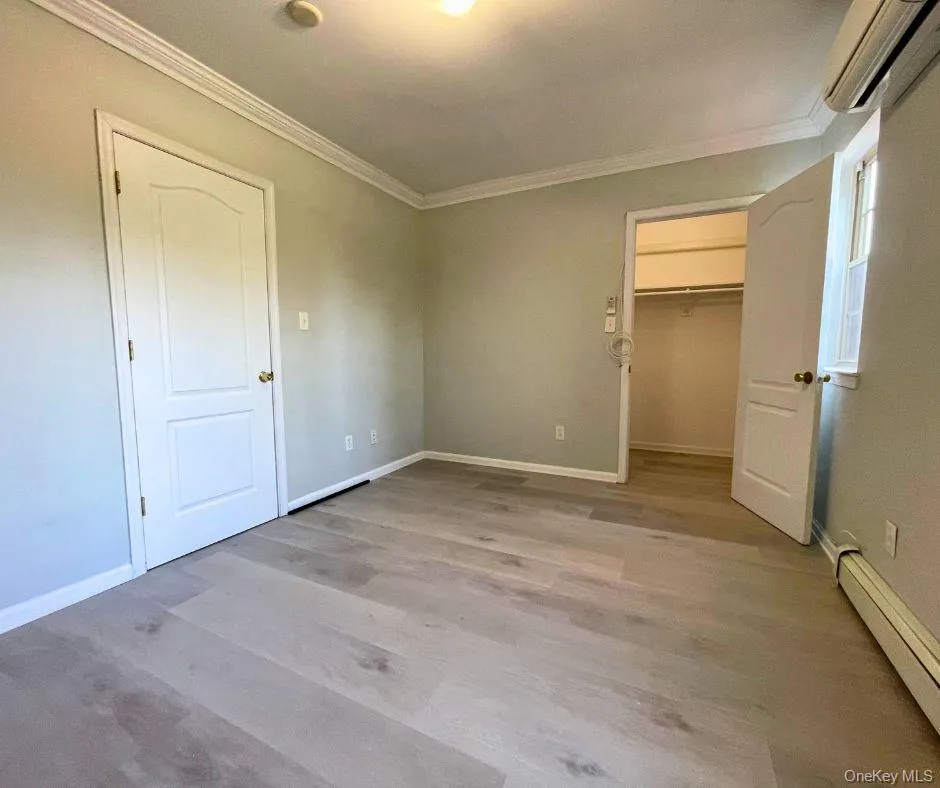Unfurnished bedroom featuring a walk in closet, baseboard heating, ornamental molding, an AC wall unit, and light wood finished floors Unfurnished bedroom featuring a walk in closet, baseboard heating, ornamental molding, an AC wall unit, and light wood finished floors