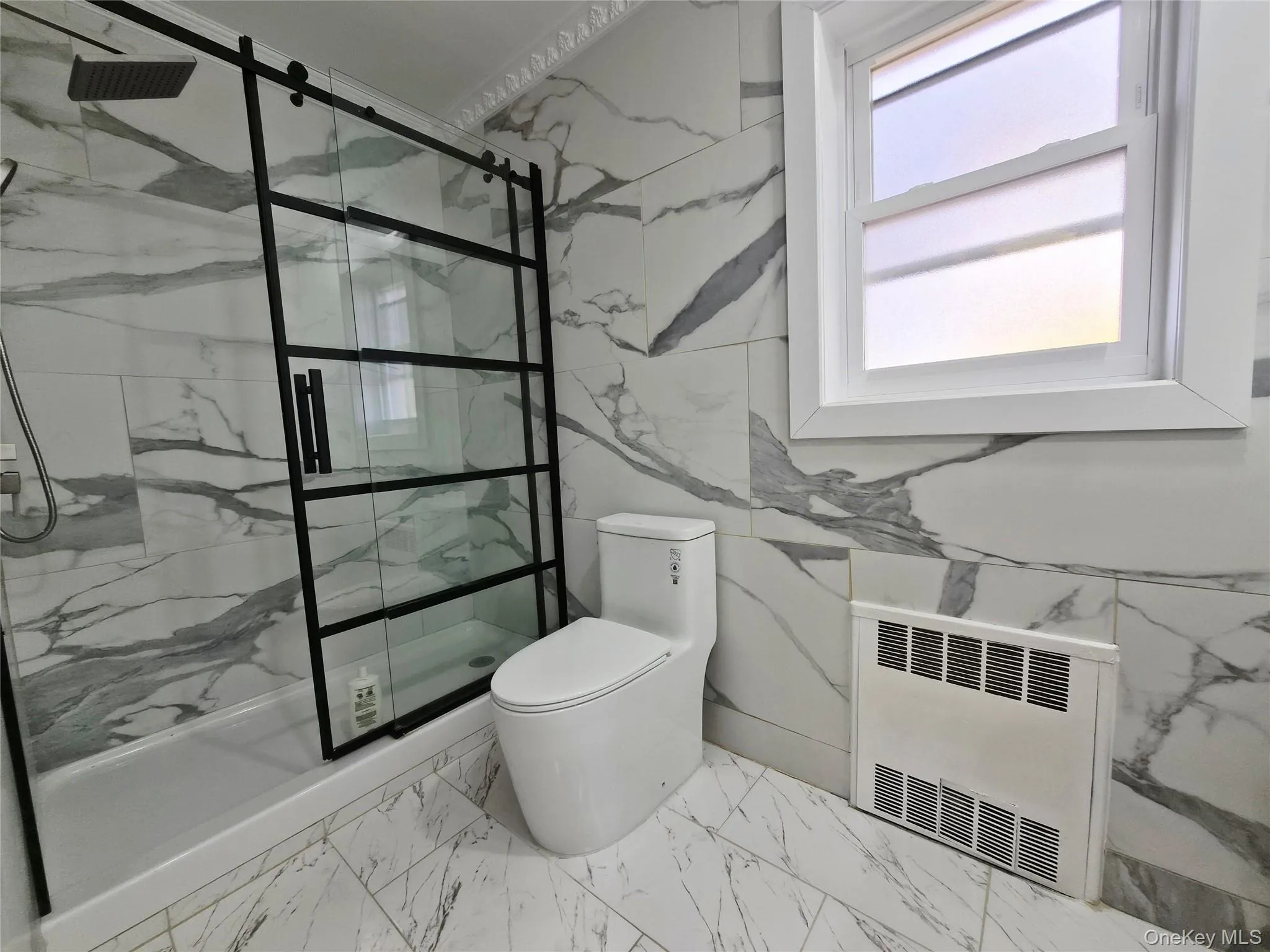 Full bathroom featuring light marble finish floors, tile walls, a marble finish shower, heating unit, and radiator Full bathroom featuring light marble finish floors, tile walls, a marble finish shower, heating unit, and radiator