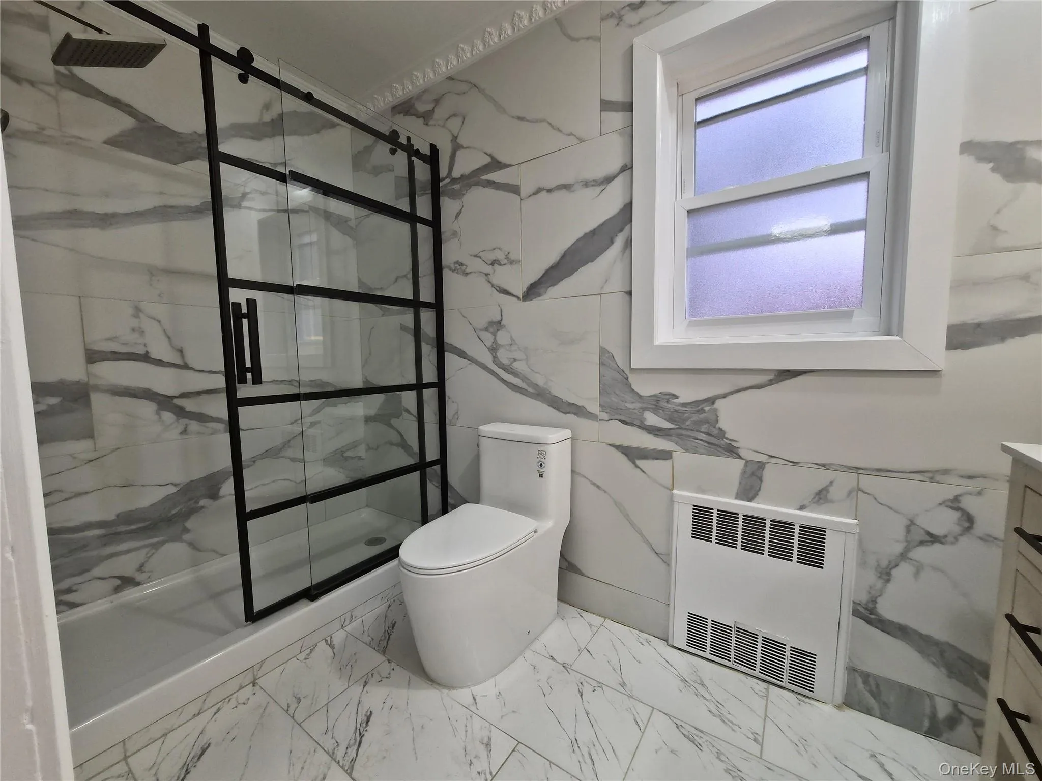 Full bath with light marble finish flooring, a marble finish shower, vanity, radiator, and tile walls Full bath with light marble finish flooring, a marble finish shower, vanity, radiator, and tile walls