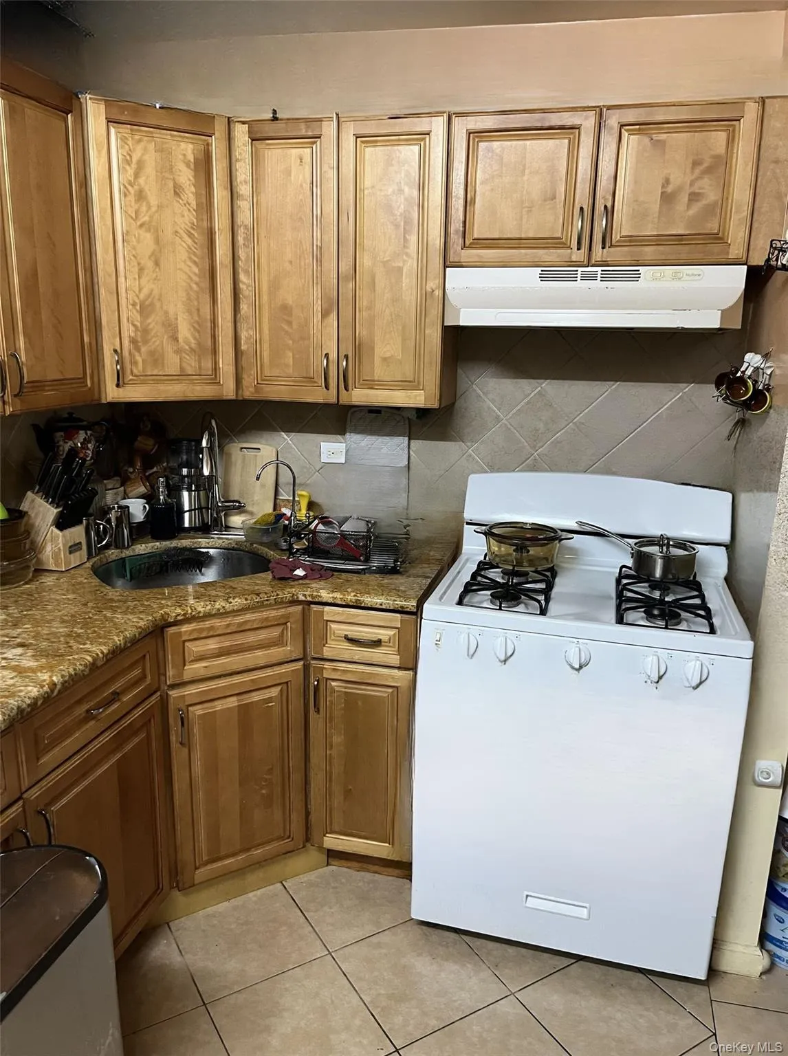 Kitchen featuring white range with gas stovetop, brown cabinetry, dark stone countertops, under cabinet range hood, and light tile patterned floors Kitchen featuring white range with gas stovetop, brown cabinetry, dark stone countertops, under cabinet range hood, and light tile patterned floors