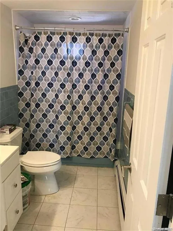 Full bath with a shower with shower curtain, light tile patterned floors, tile walls, vanity, and a baseboard radiator Full bath with a shower with shower curtain, light tile patterned floors, tile walls, vanity, and a baseboard radiator