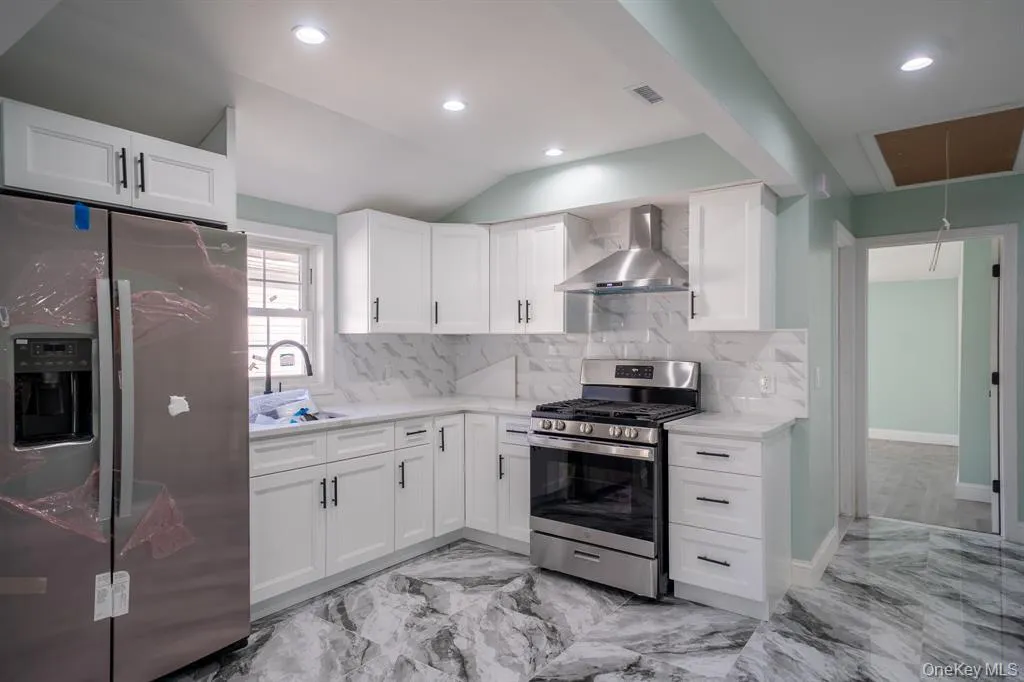 Kitchen featuring stainless steel appliances, white cabinetry, wall chimney exhaust hood, light marble finish floors, and vaulted ceiling Kitchen featuring stainless steel appliances, white cabinetry, wall chimney exhaust hood, light marble finish floors, and vaulted ceiling