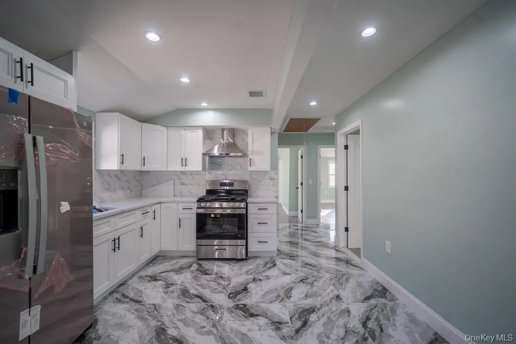 Kitchen featuring stainless steel appliances, white cabinets, decorative backsplash, vaulted ceiling, and wall chimney range hood Kitchen featuring stainless steel appliances, white cabinets, decorative backsplash, vaulted ceiling, and wall chimney range hood