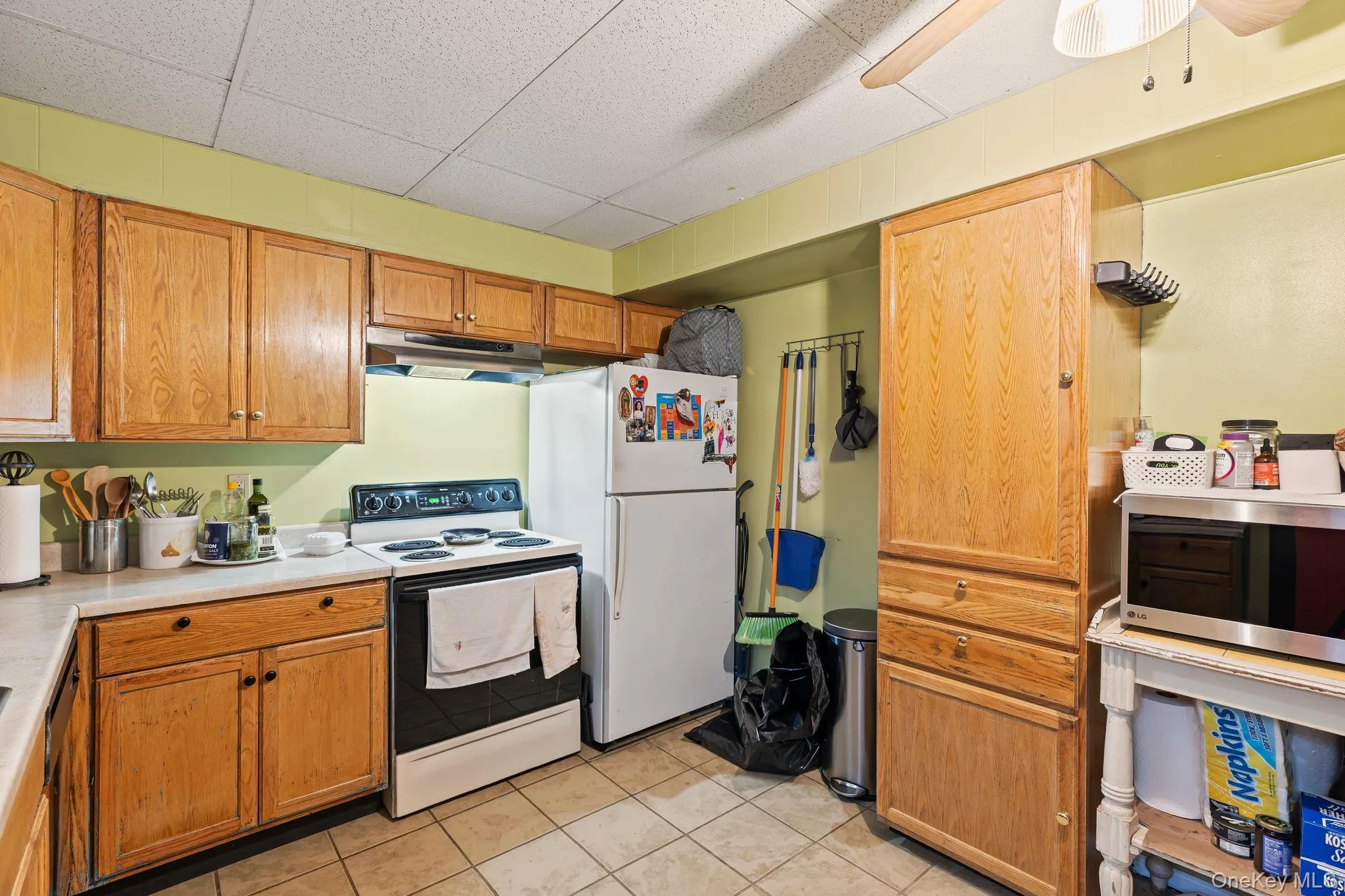 Kitchen featuring white appliances, light countertops, under cabinet range hood, brown cabinetry, and light tile patterned floors Kitchen featuring white appliances, light countertops, under cabinet range hood, brown cabinetry, and light tile patterned floors