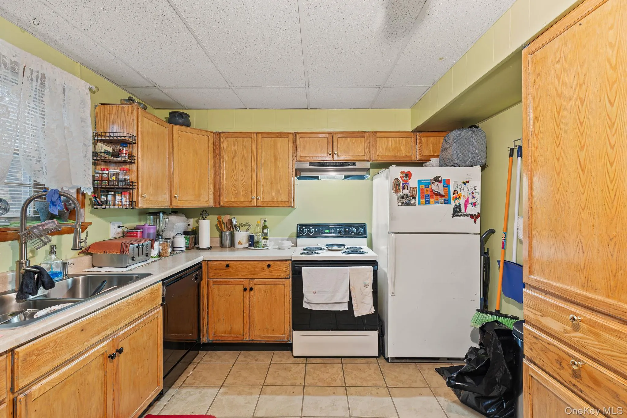 Kitchen with white appliances, a drop ceiling, light countertops, under cabinet range hood, and light tile patterned floors Kitchen with white appliances, a drop ceiling, light countertops, under cabinet range hood, and light tile patterned floors
