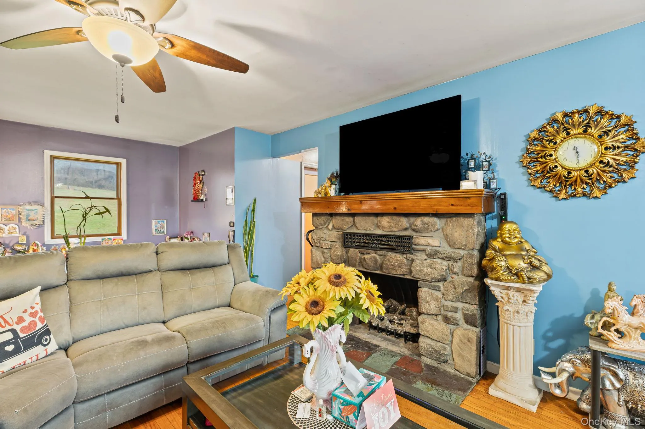 Living room featuring wood finished floors, a stone fireplace, and ceiling fan Living room featuring wood finished floors, a stone fireplace, and ceiling fan