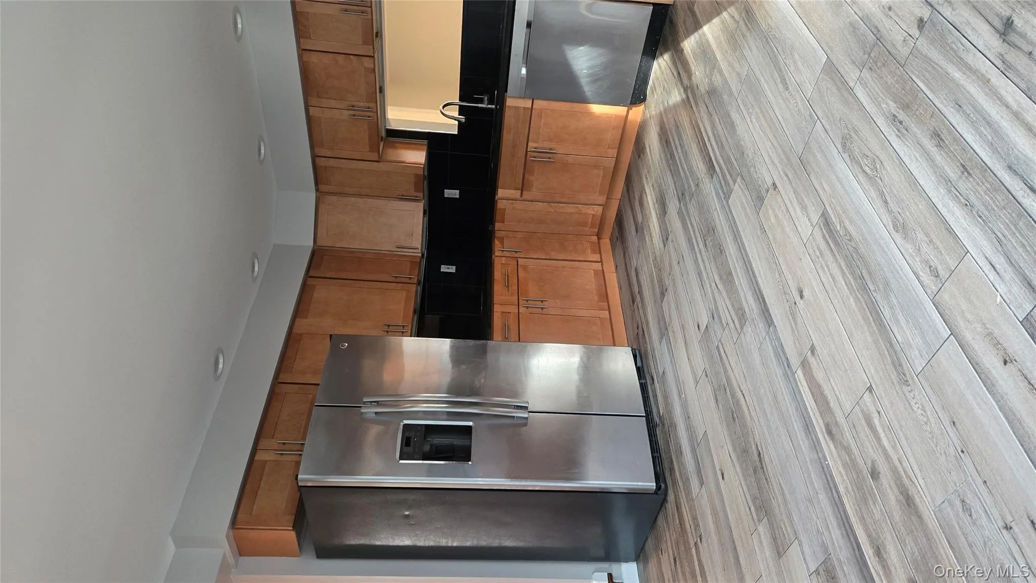 Kitchen featuring stainless steel fridge, dishwasher, dark countertops, light wood-type flooring, and brown cabinetry Kitchen featuring stainless steel fridge, dishwasher, dark countertops, light wood-type flooring, and brown cabinetry