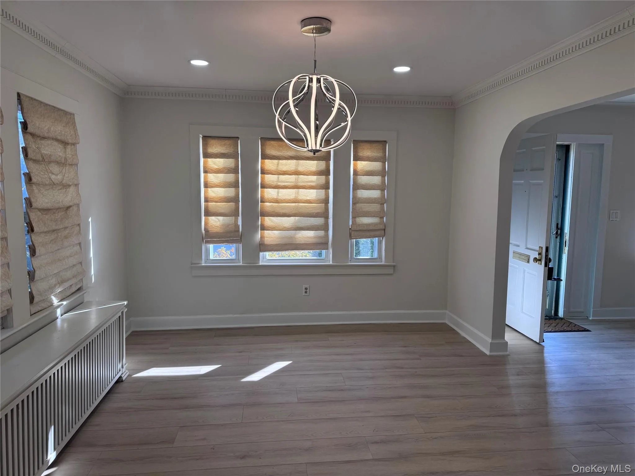 Unfurnished dining area featuring radiator heating unit, arched walkways, crown molding, a chandelier, and light wood finished floors Unfurnished dining area featuring radiator heating unit, arched walkways, crown molding, a chandelier, and light wood finished floors