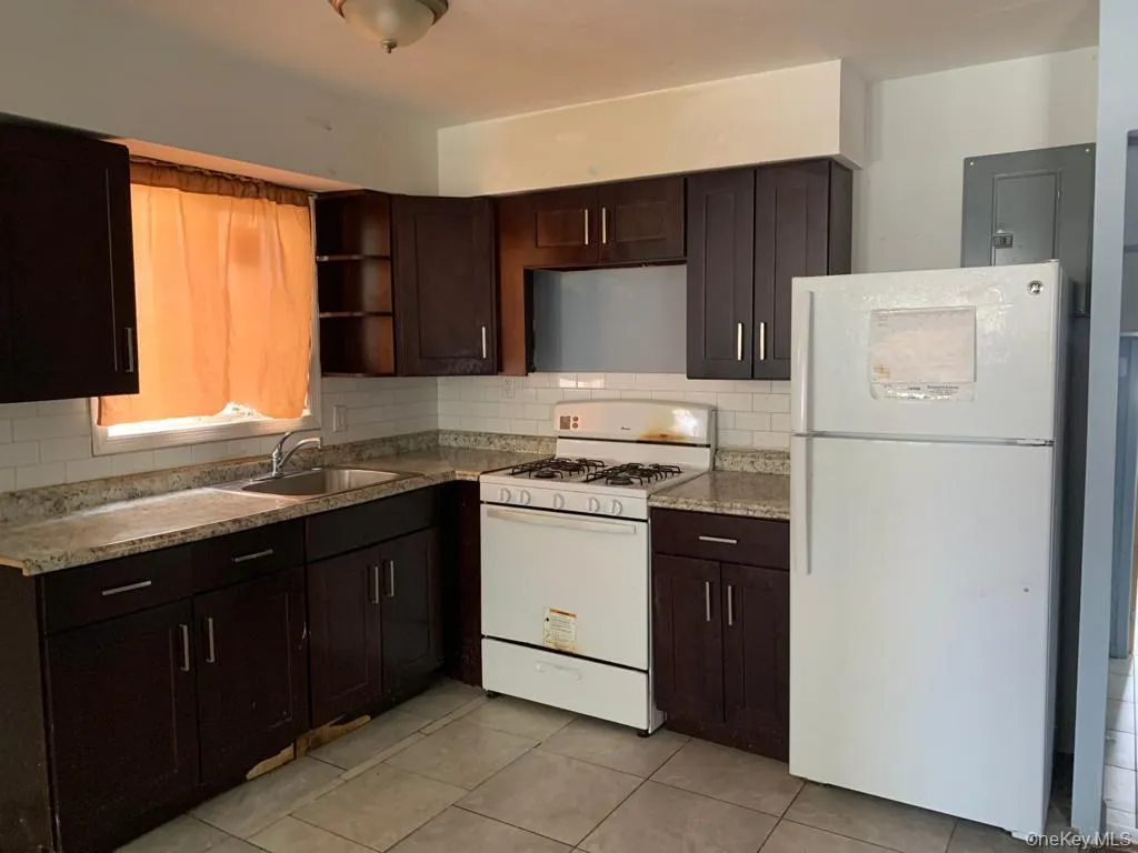 Kitchen featuring dark brown cabinetry, white appliances, decorative backsplash, light tile patterned floors, and electric panel Kitchen featuring dark brown cabinetry, white appliances, decorative backsplash, light tile patterned floors, and electric panel
