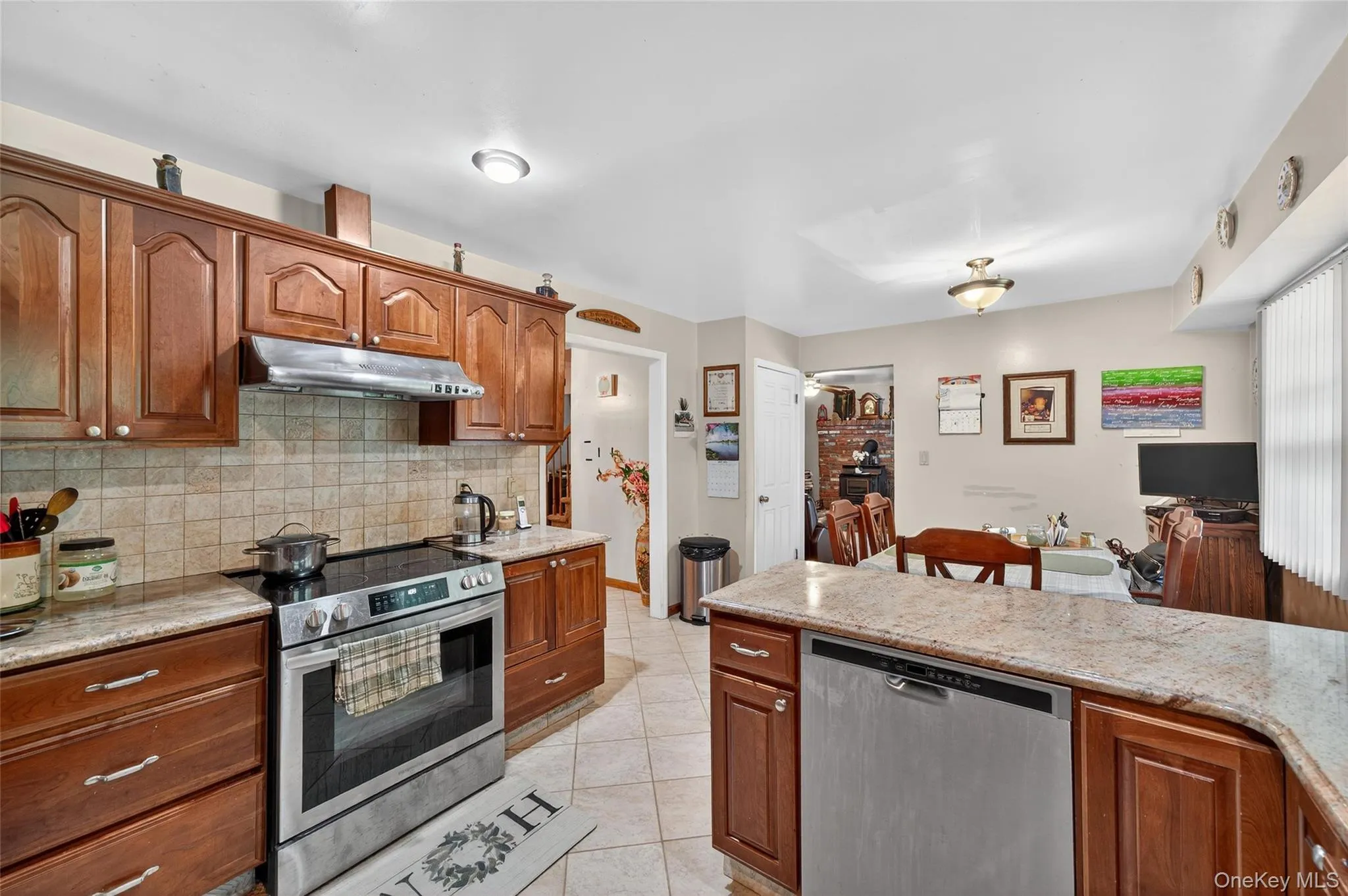 Kitchen featuring appliances with stainless steel finishes, brown cabinets, light tile patterned floors, under cabinet range hood, and decorative backsplash Kitchen featuring appliances with stainless steel finishes, brown cabinets, light tile patterned floors, under cabinet range hood, and decorative backsplash