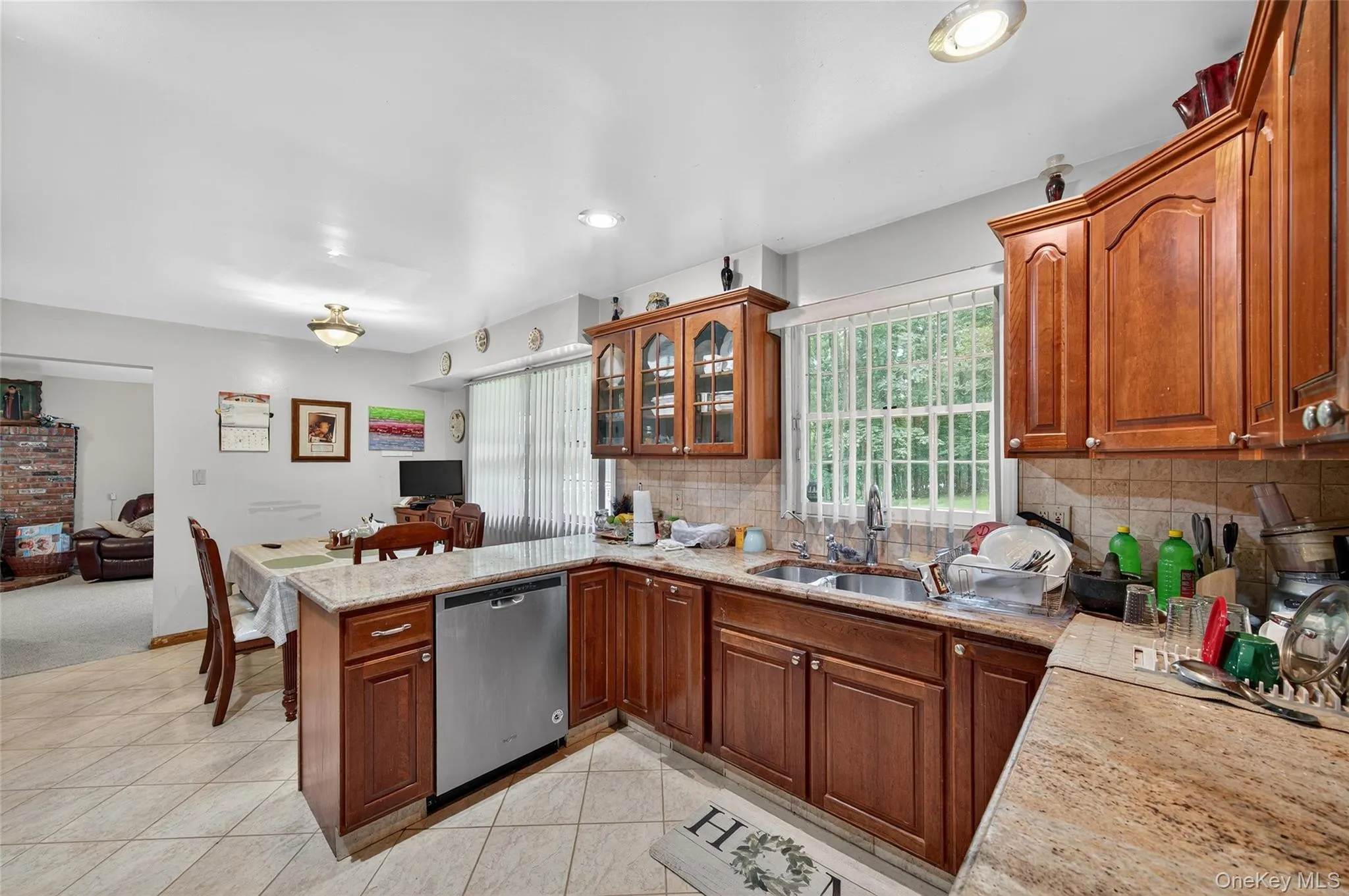 Kitchen featuring stainless steel dishwasher, glass insert cabinets, brown cabinets, tasteful backsplash, and open floor plan Kitchen featuring stainless steel dishwasher, glass insert cabinets, brown cabinets, tasteful backsplash, and open floor plan