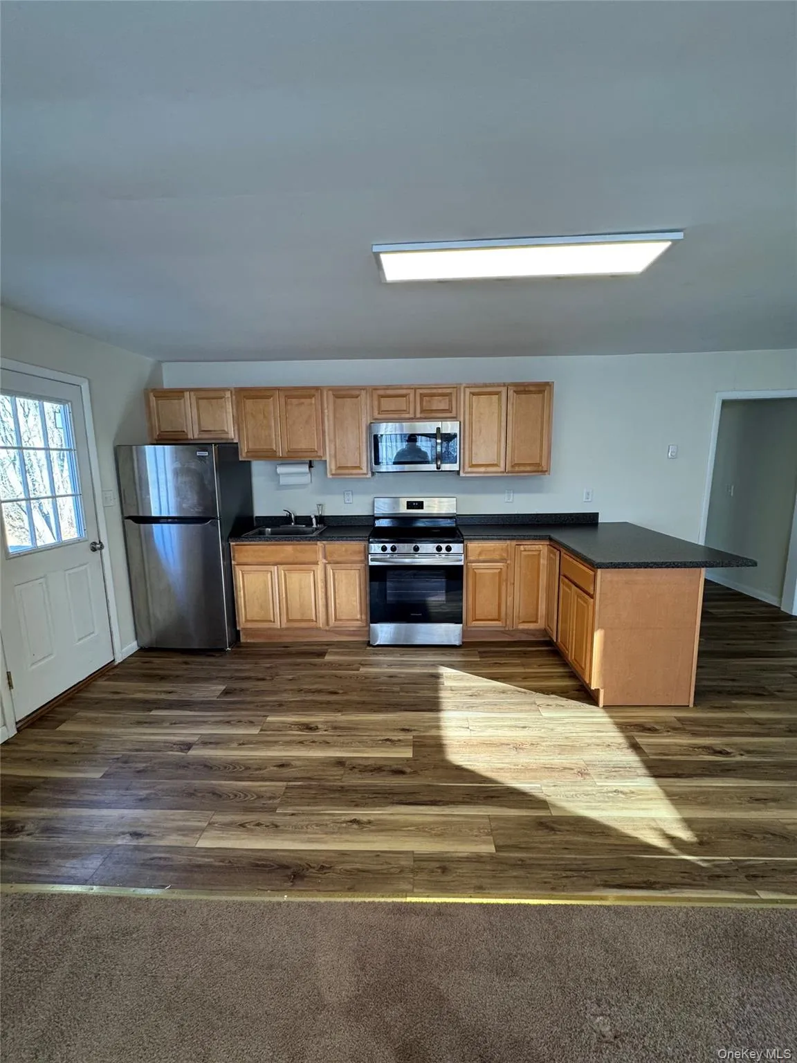 Kitchen with dark countertops, appliances with stainless steel finishes, a peninsula, dark wood-style floors, and brown cabinetry Kitchen with dark countertops, appliances with stainless steel finishes, a peninsula, dark wood-style floors, and brown cabinetry