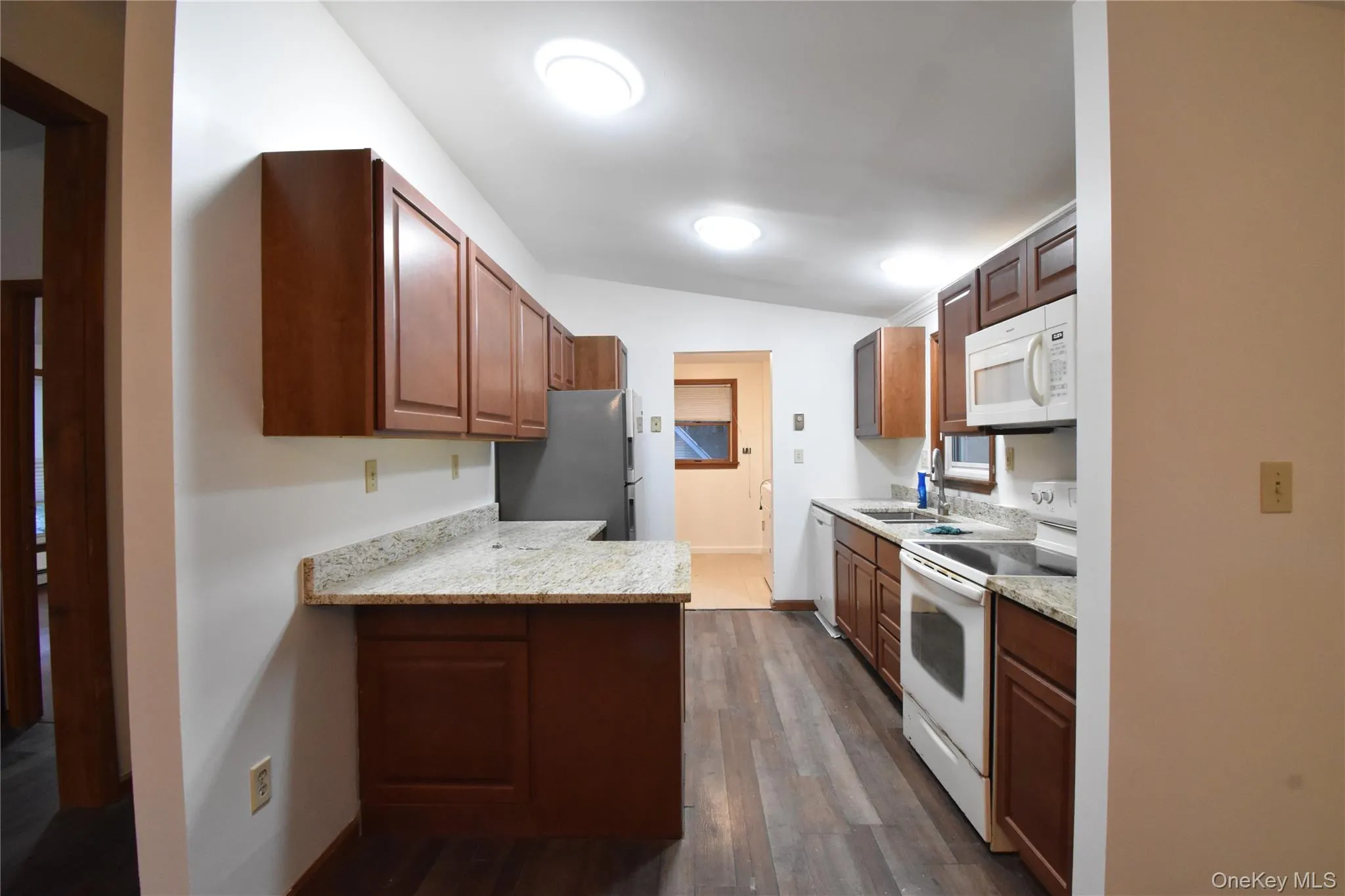 Kitchen featuring white appliances, dark wood-style flooring, vaulted ceiling, light stone countertops, and a peninsula Kitchen featuring white appliances, dark wood-style flooring, vaulted ceiling, light stone countertops, and a peninsula