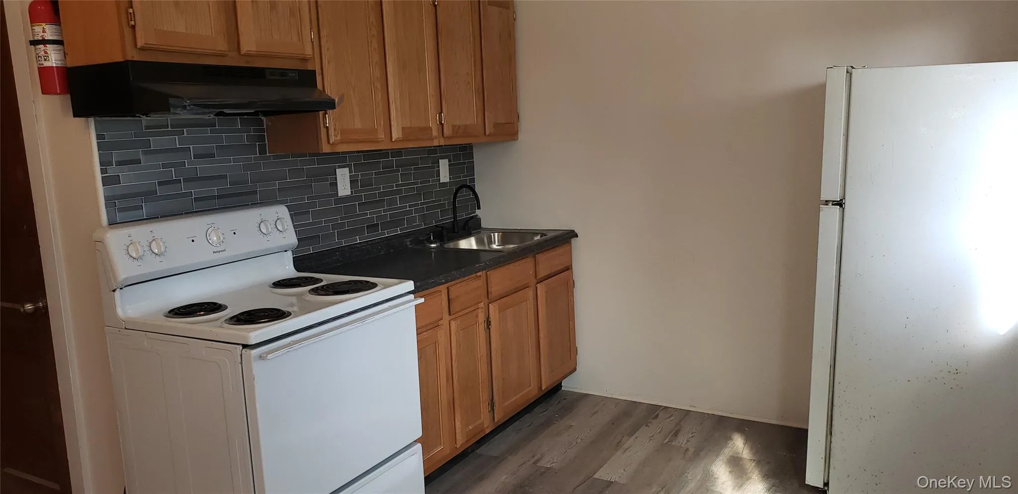 Kitchen featuring white appliances, decorative backsplash, brown cabinetry, under cabinet range hood, and dark wood finished floors Kitchen featuring white appliances, decorative backsplash, brown cabinetry, under cabinet range hood, and dark wood finished floors