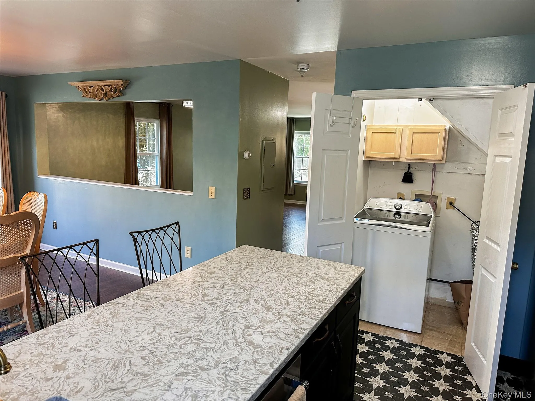 Kitchen featuring dark cabinetry, washer / dryer, light countertops, light brown cabinetry, and electric panel Kitchen featuring dark cabinetry, washer / dryer, light countertops, light brown cabinetry, and electric panel