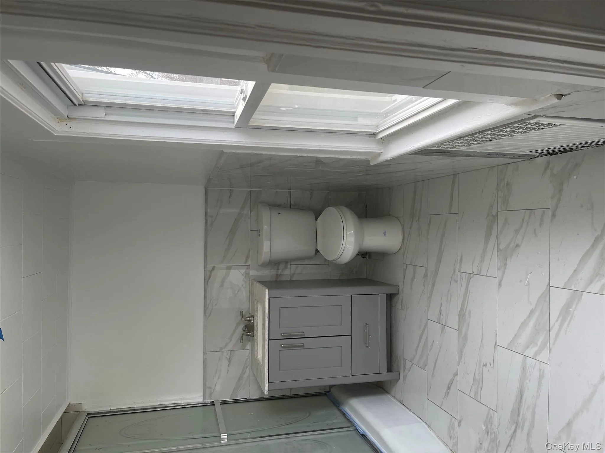 Bathroom with vanity, wainscoting, bath / shower combo with glass door, tile walls, and light marble finish floors Bathroom with vanity, wainscoting, bath / shower combo with glass door, tile walls, and light marble finish floors